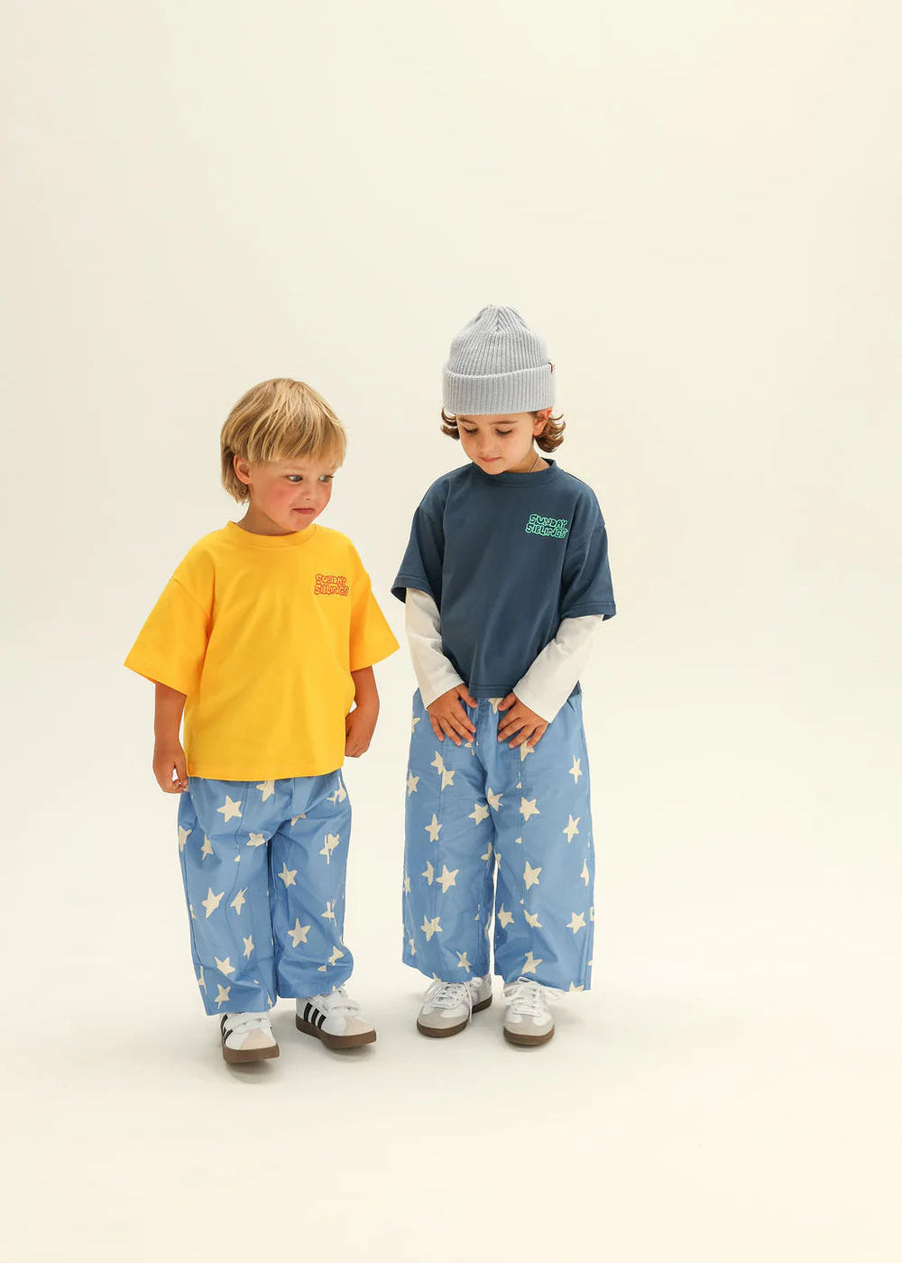 Two children wearing matching outfits with blue pants and colorful shirts on a beige background