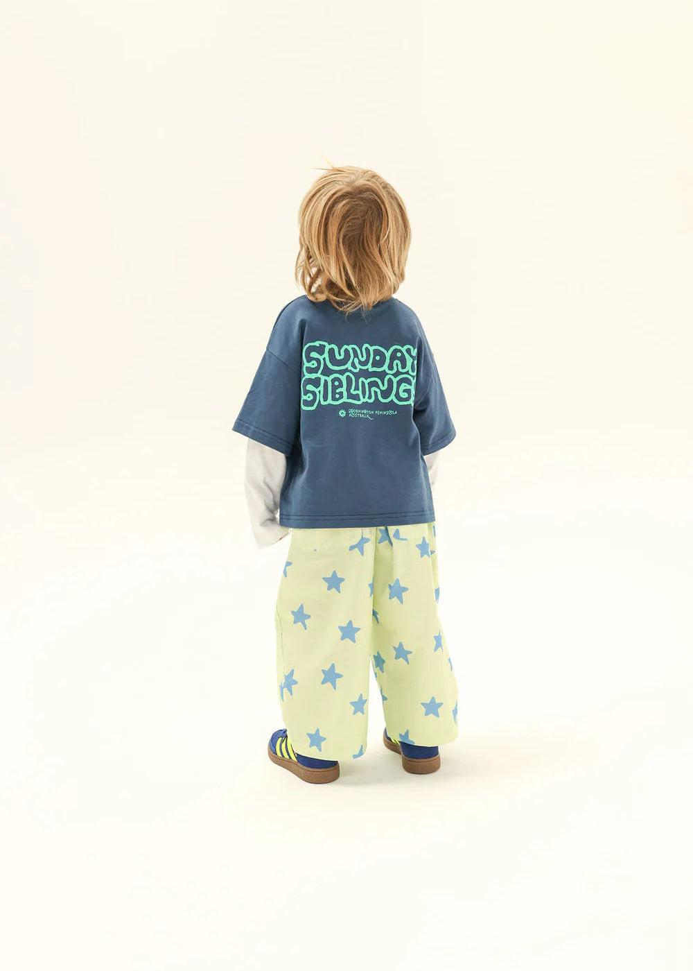 Child wearing a blue shirt with 'Sundaysibling' text and light-colored pants with star patterns on a white background