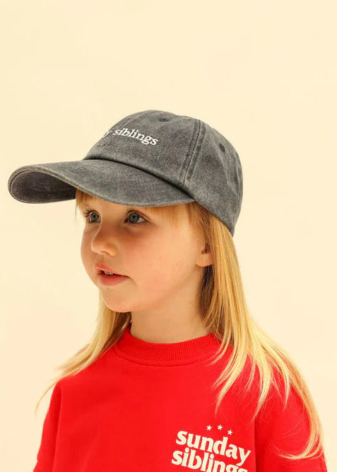 Child wearing a gray cap and red shirt with 'sunday siblings' branding on a beige background