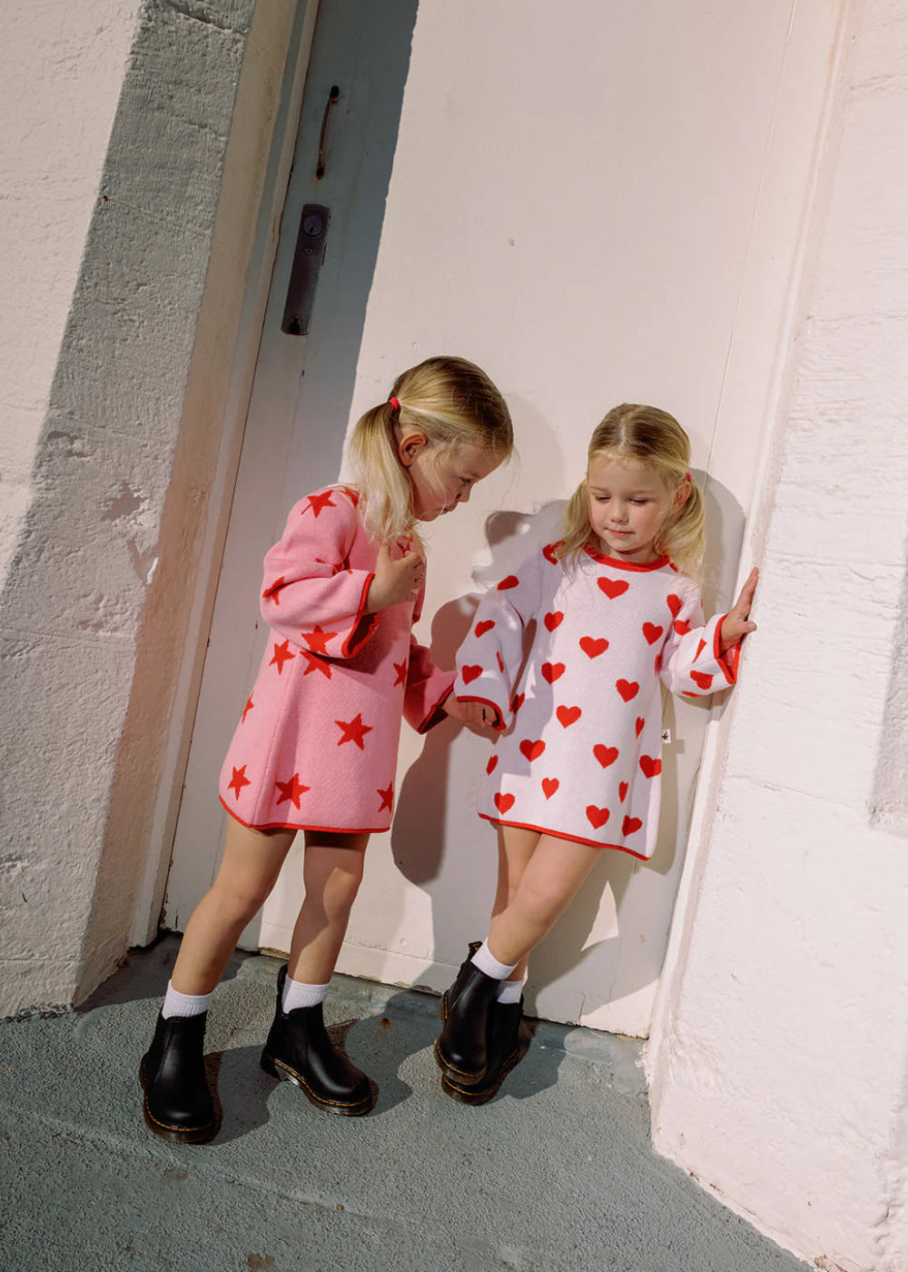 Two children in matching outfits with star and heart patterns standing against a white wall.