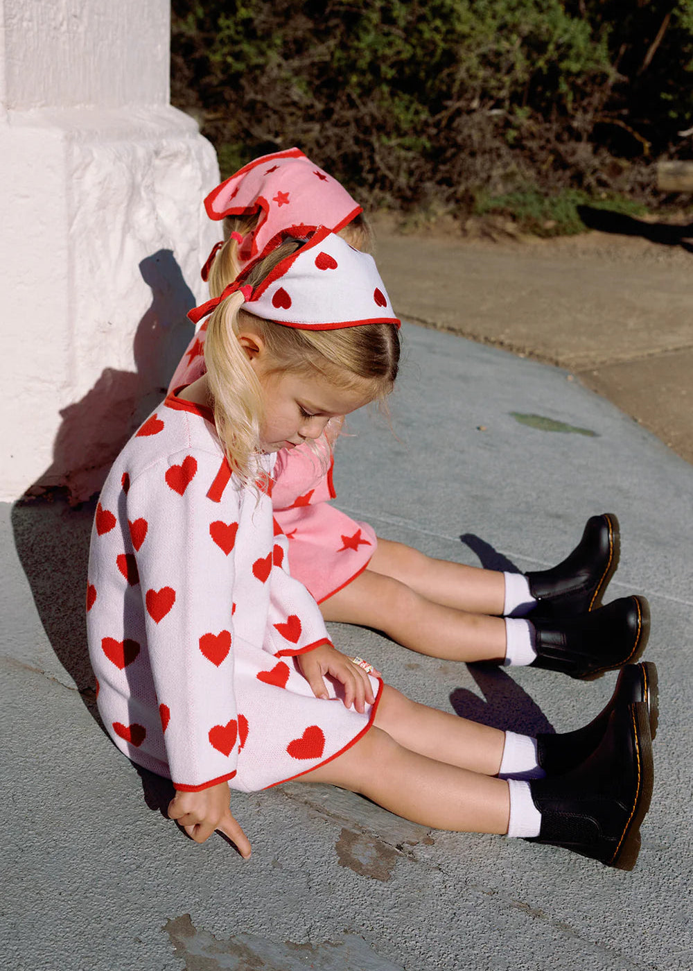 Child sitting on a sidewalk wearing a heart-patterned dress and red headband.
