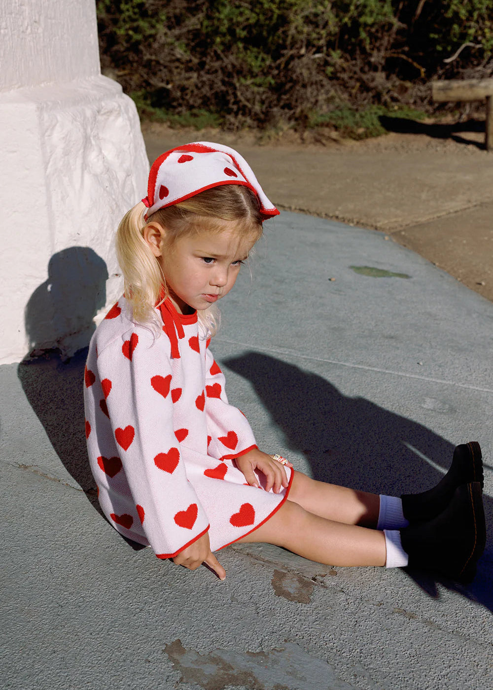 Young girl sitting on a stone surface wearing a red and white dress with heart patterns.