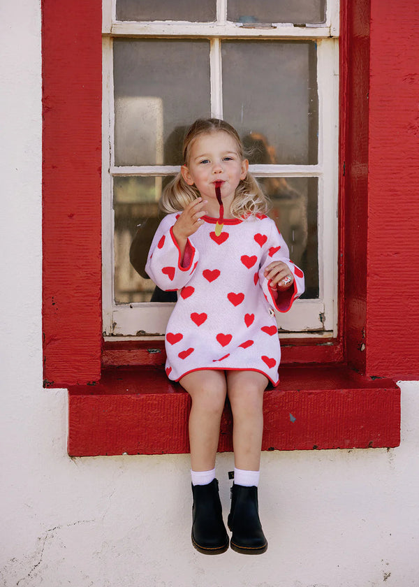Child wearing a red dress with heart patterns sitting in front of a red window frame.