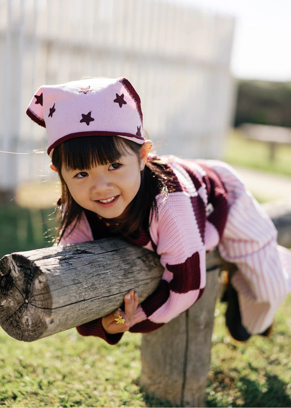 Child wearing a pink hat with star patterns and a patterned sweater, leaning on a wooden post outdoors.