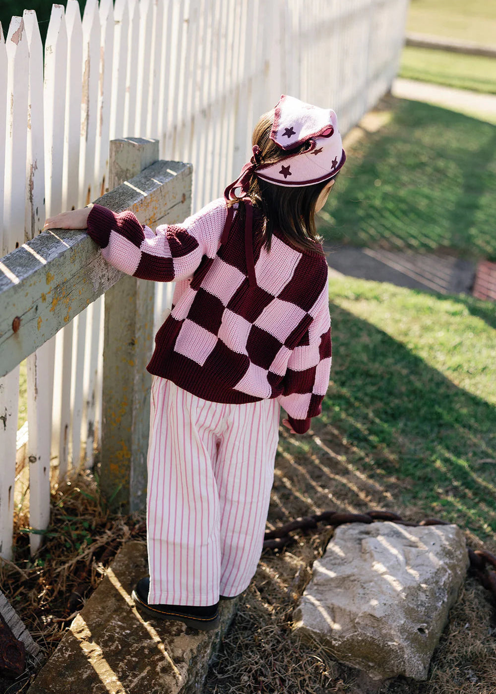 Person wearing a checkered sweater and striped pants leaning against a white picket fence.