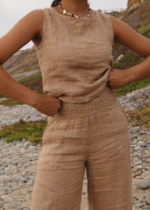 Person wearing a beige sleeveless top and pants standing on a rocky beach.
