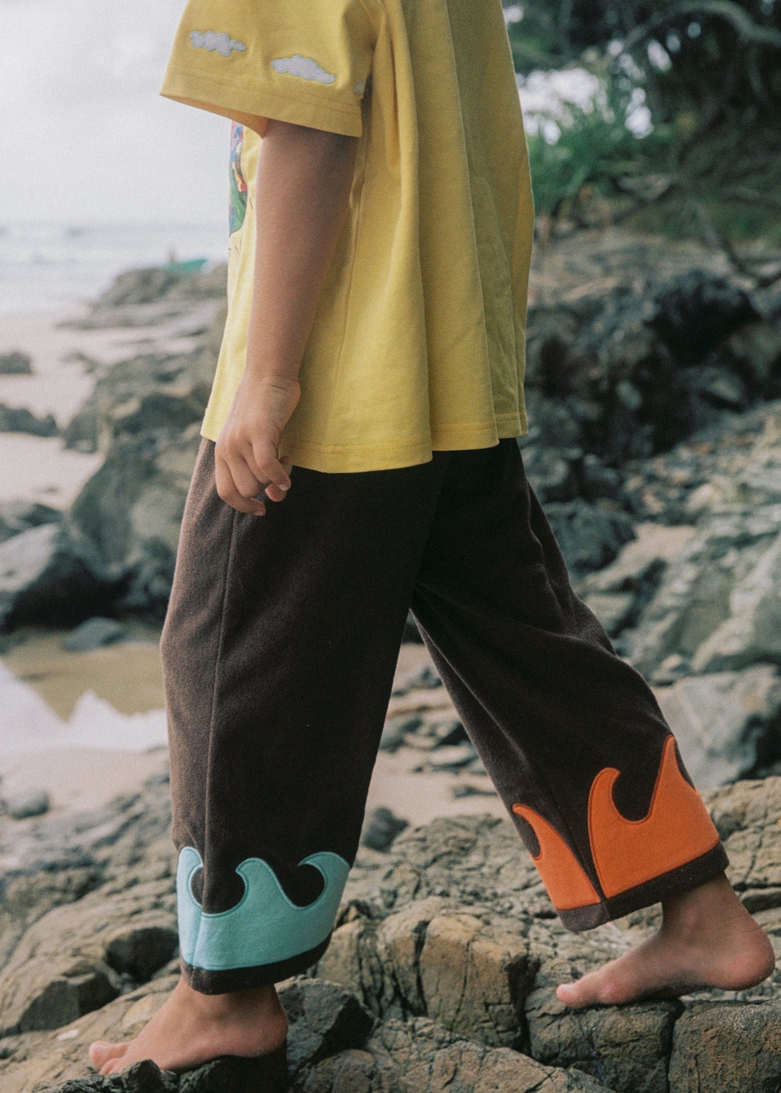 Person wearing a yellow shirt and brown pants with orange and blue accents on a rocky beach.