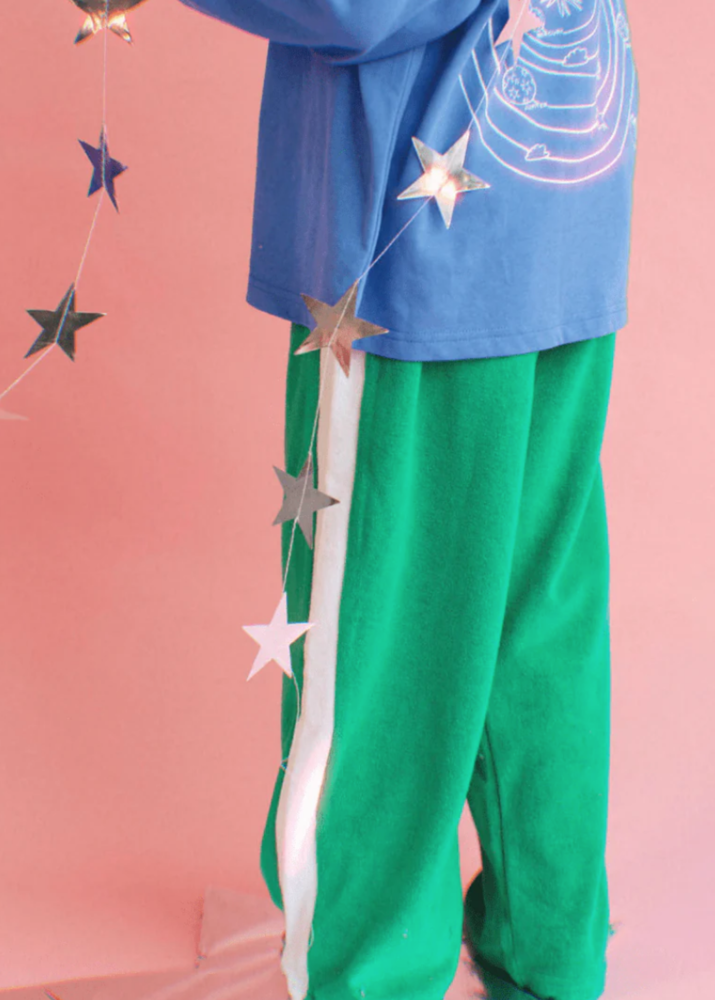 Person wearing a blue top with star designs and green pants on a pink background