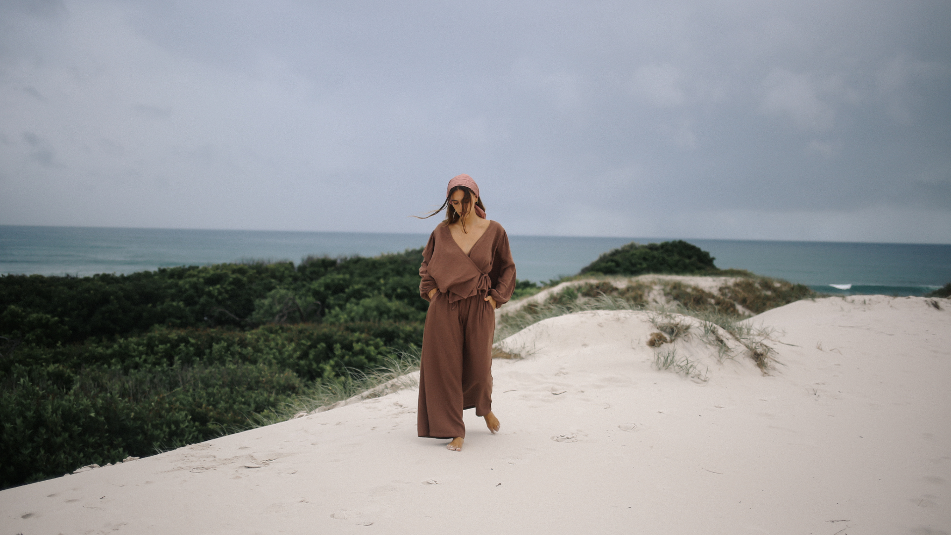 Woman in a brown jumpsuit standing on a sandy dune with ocean and greenery in the background