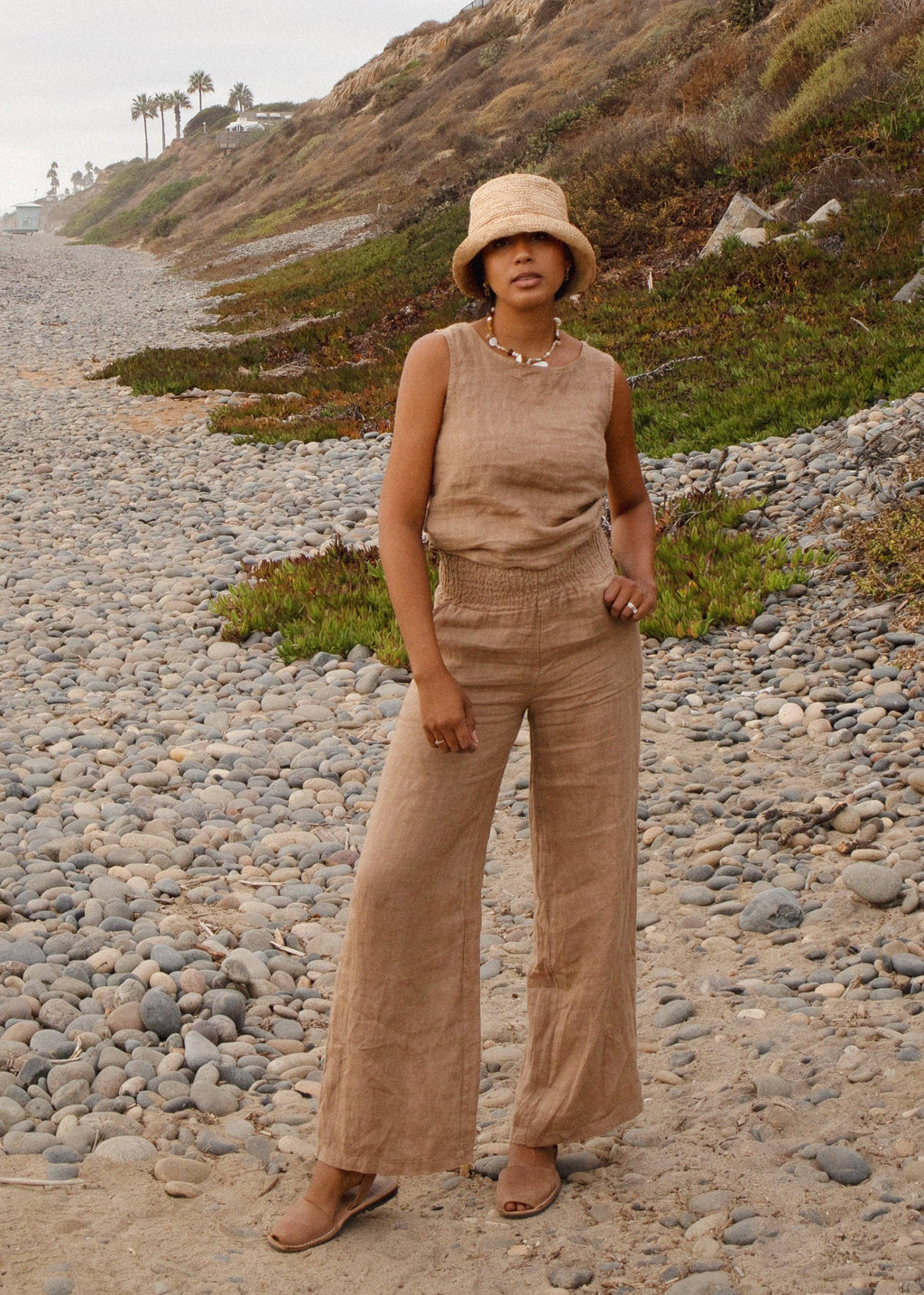 Woman in beige outfit standing on a rocky beach with grassy hillside in the background