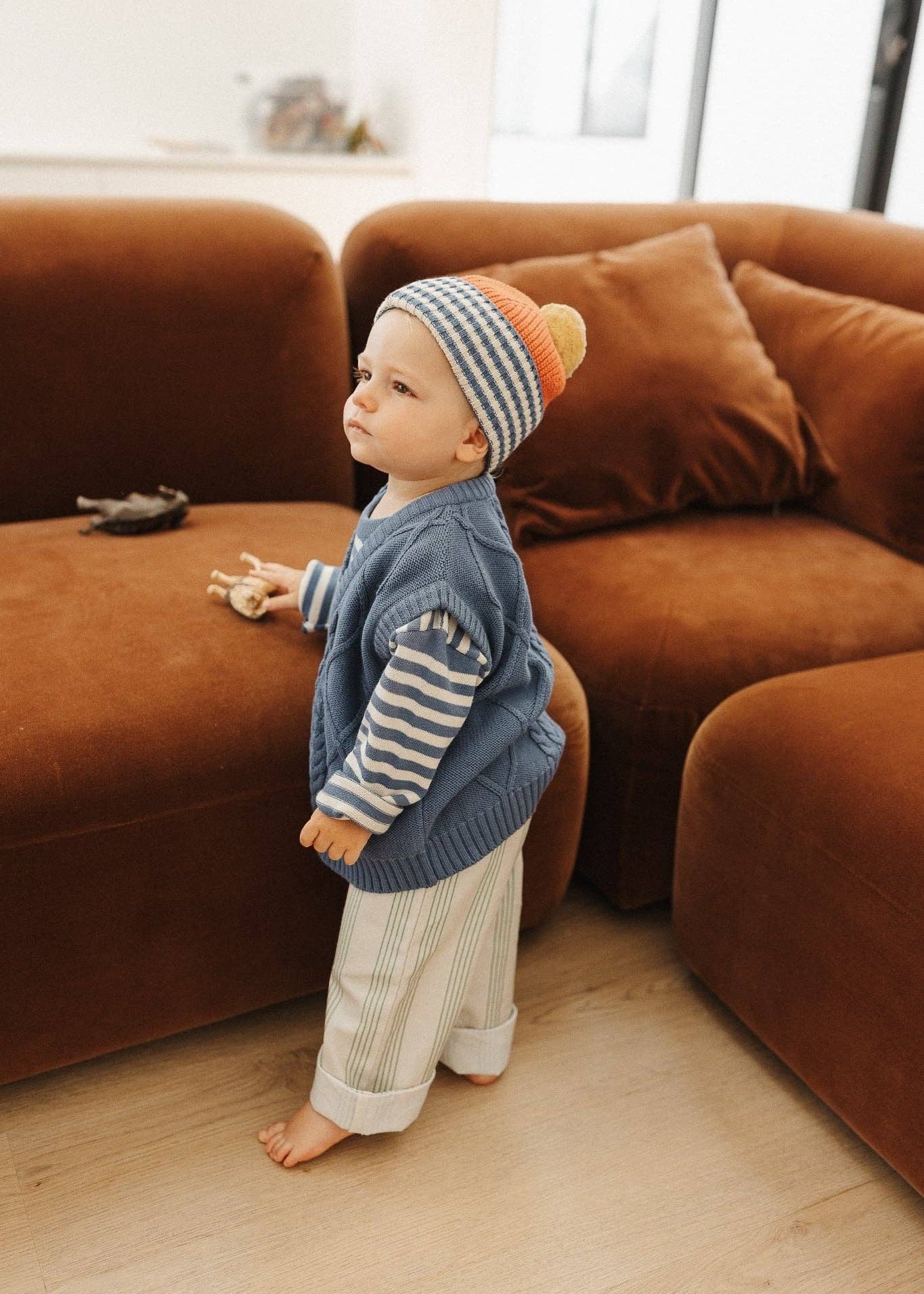 Child wearing a striped hat and blue sweater standing in front of a brown sofa.