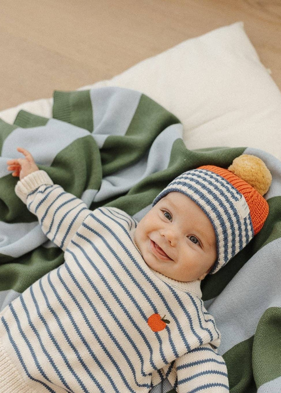 Baby lying on a blanket with a striped hat and orange socks on a wooden floor.