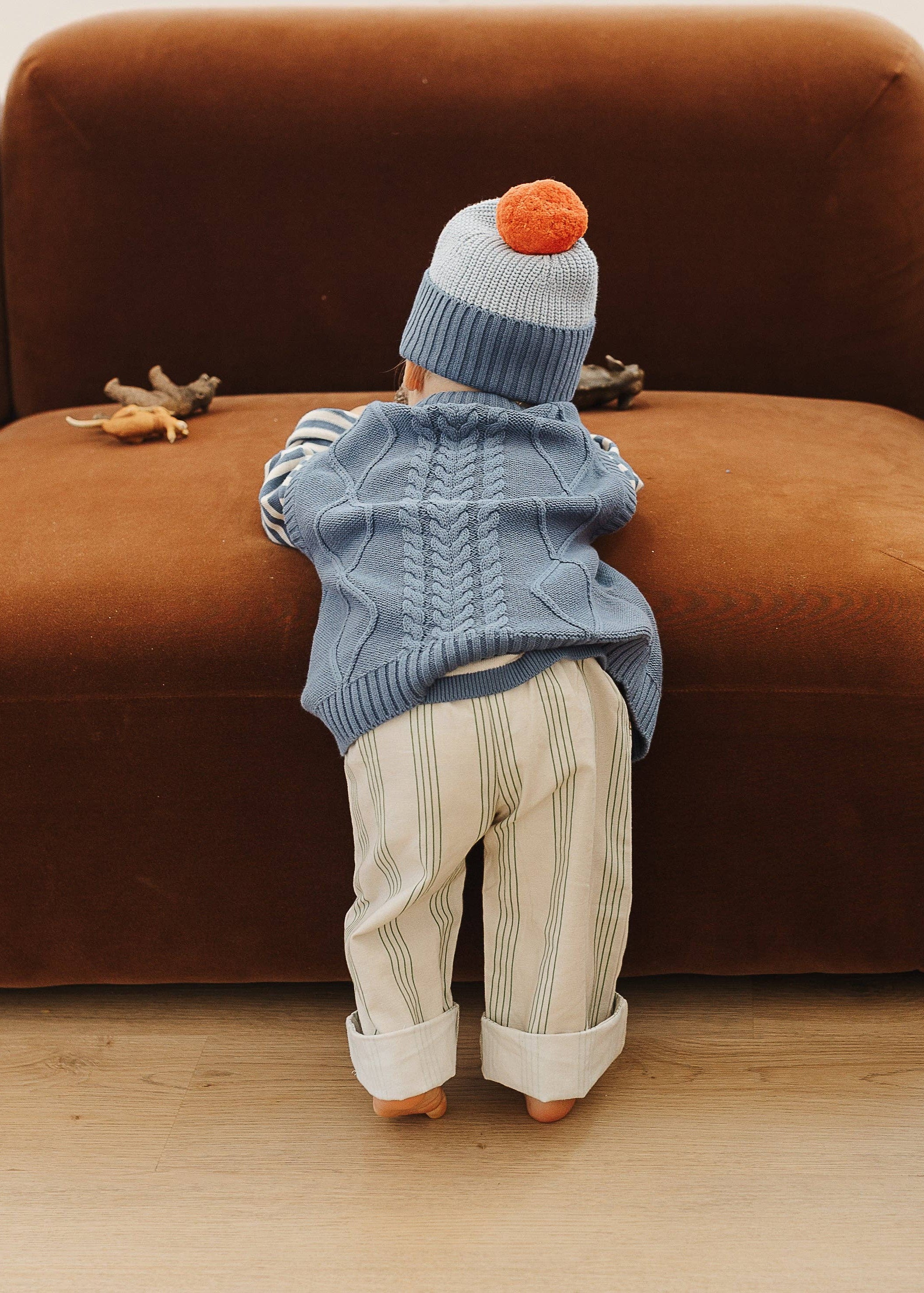 Child wearing a blue sweater and striped pants standing on a wooden floor next to a brown sofa.