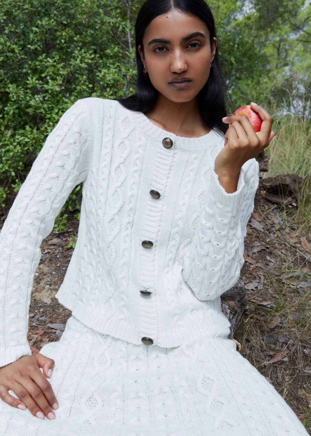 Woman in a white cable knit cardigan holding an apple outdoors.