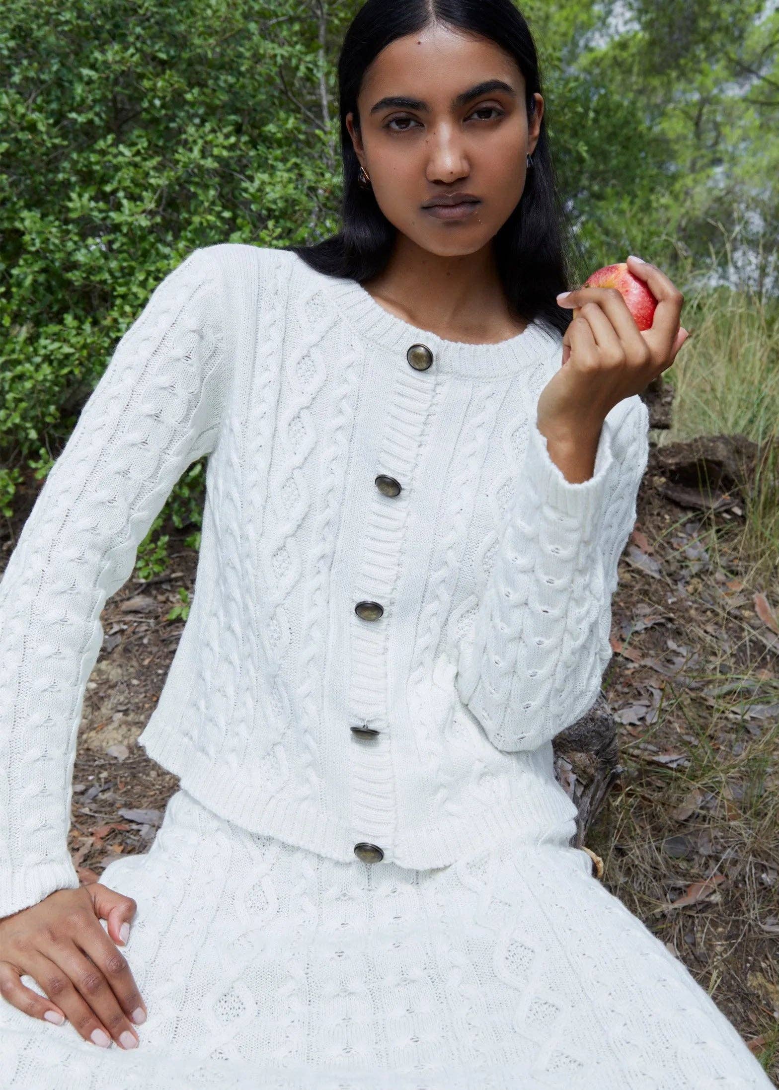 Woman in a white cable knit cardigan holding an apple outdoors.