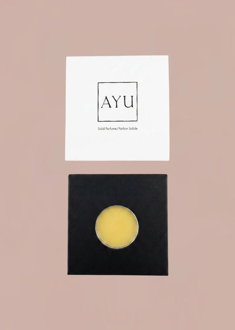 A round yellow solid facial balm in a clear container, displayed on a black square card below a white card printed with the brand name 'AYU' on a solid dusty pink background.