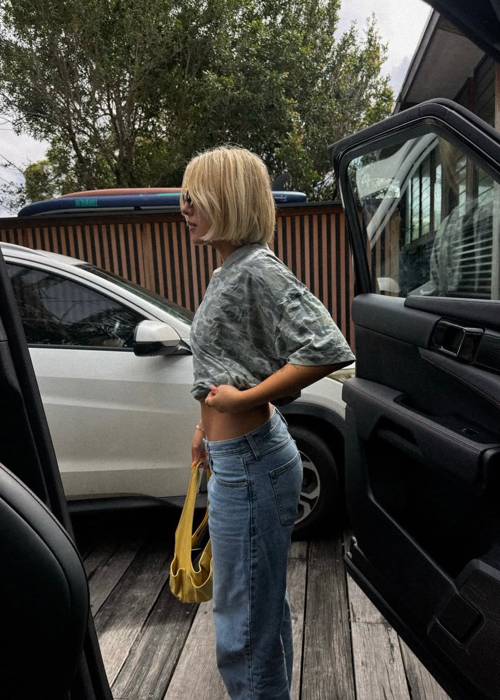A woman with a blonde bob stands between open cars on a wooden deck, wearing a light gray patterned short-sleeve top and light blue jeans, holding a yellow bag.