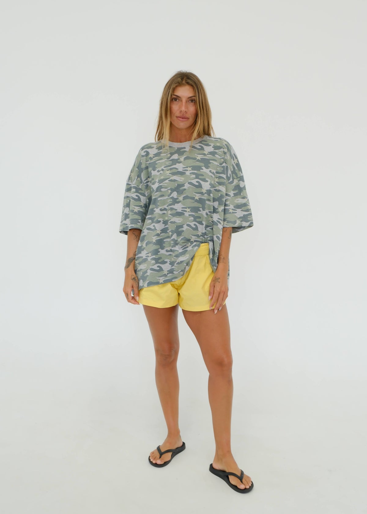 A woman modeling a loose-fit camouflage print t-shirt in muted green and gray tones, paired with bright yellow shorts and black flip-flops, standing against a seamless white studio background.