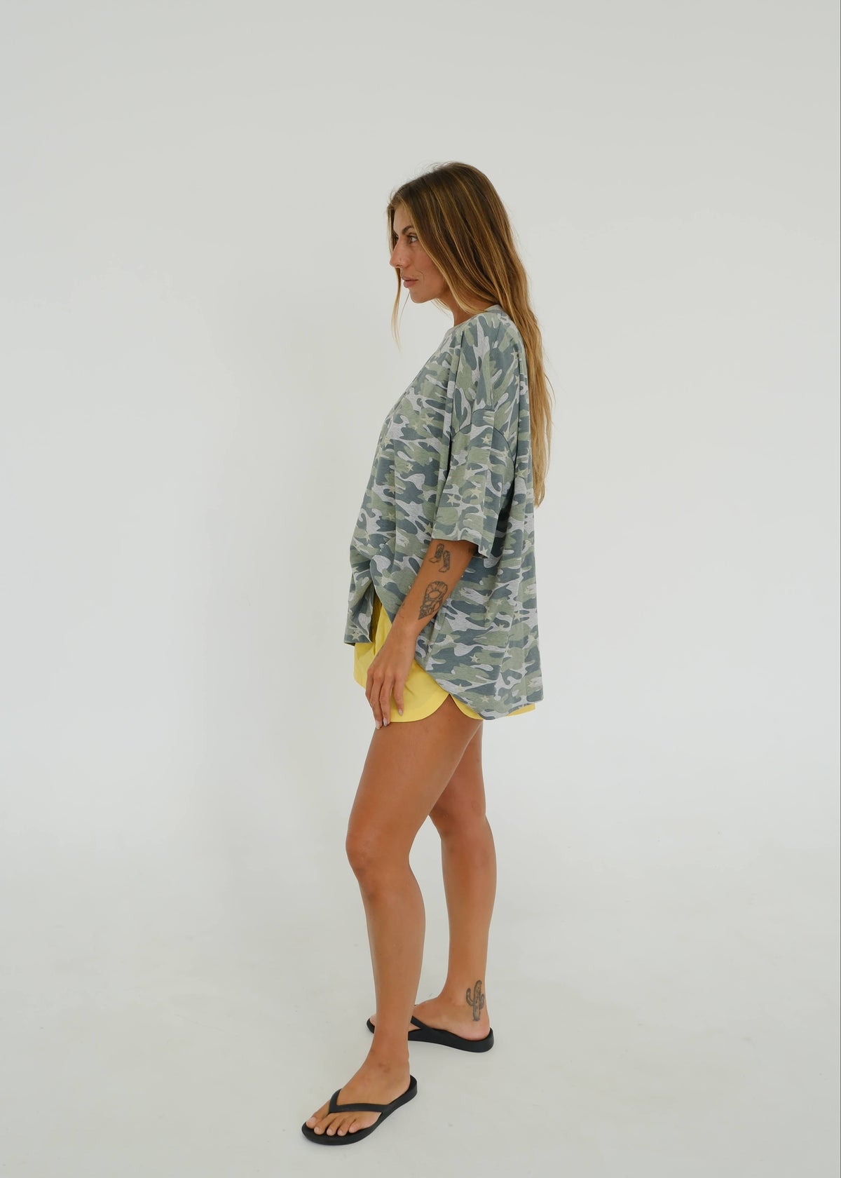 A woman stands in profile wearing a loose, oversized short-sleeve top with a blue and green camouflage pattern, paired with yellow shorts and black flip-flops, against a plain light gray studio background.