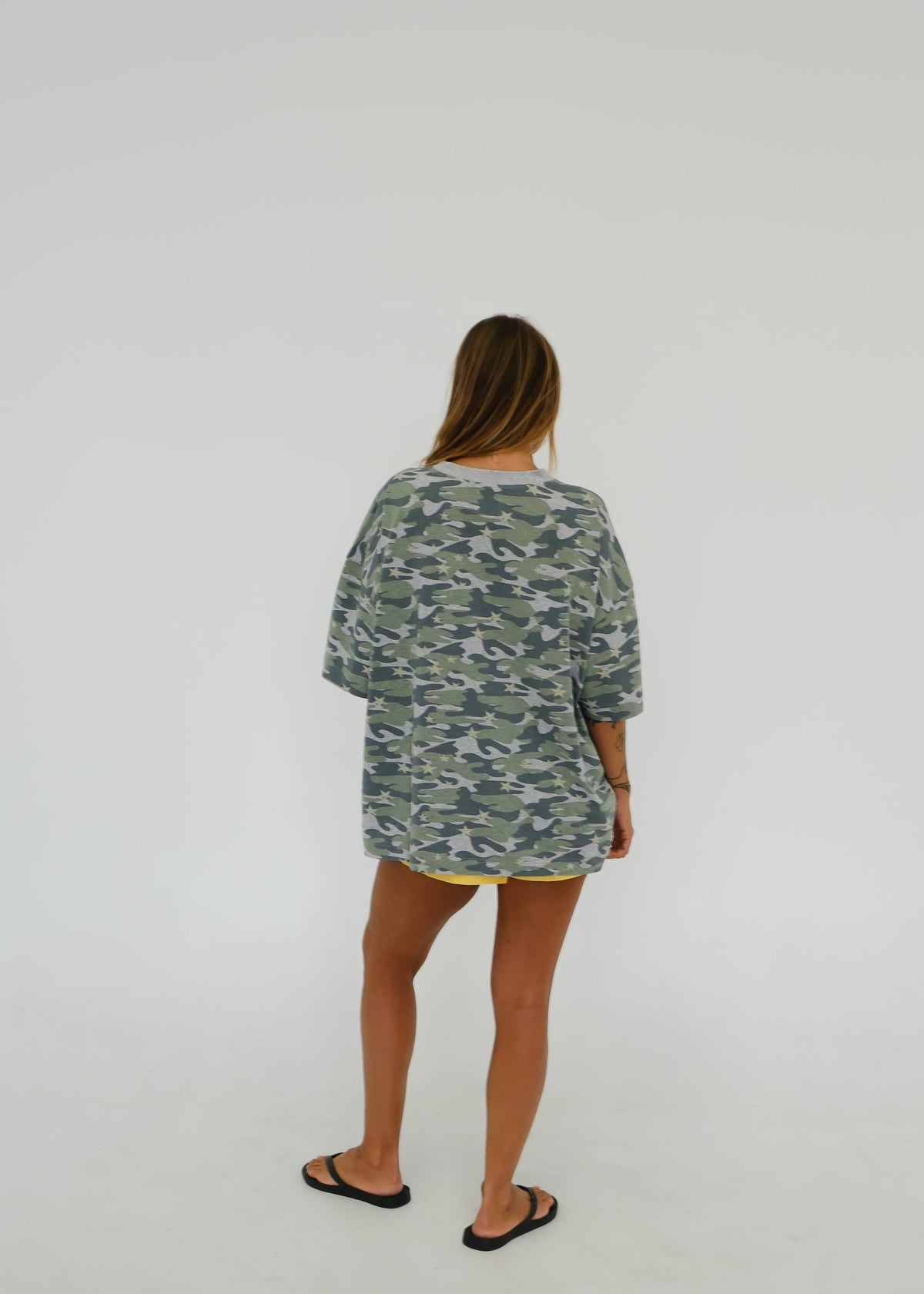 A woman viewed from behind wearing an oversized short-sleeve t-shirt with a green and gray camouflage pattern, paired with yellow trim and black flip-flops, against a plain white studio background.