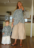 A woman and a young child wearing matching camouflage long-sleeve top and wide-leg bottoms, standing indoors in a cafe-like environment with wood floors and a counter in the background.
