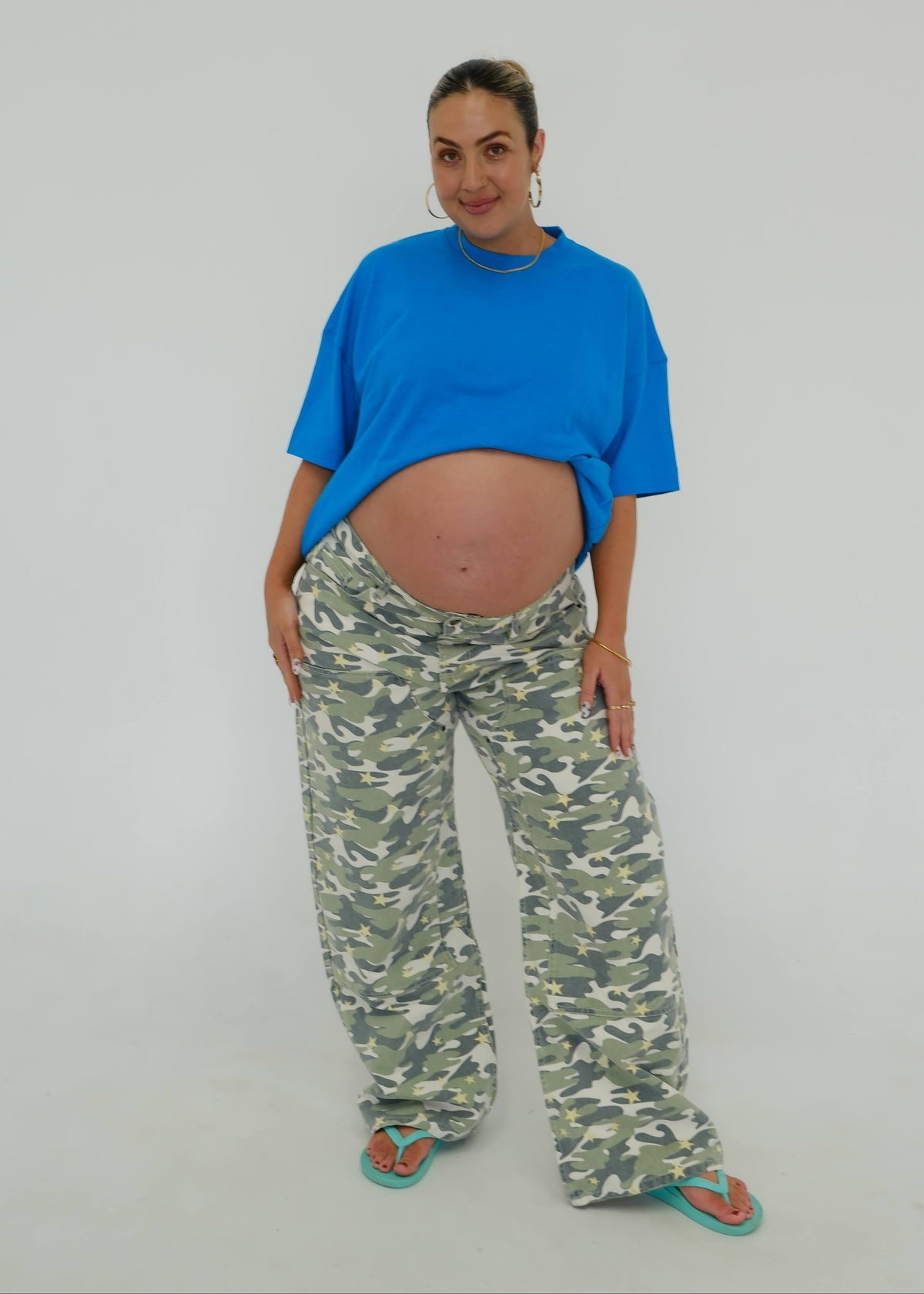 A pregnant woman posing in a studio setting, wearing an oversized bright blue t-shirt, green and white camouflage wide-leg pants, and teal flip-flops against a light gray background.