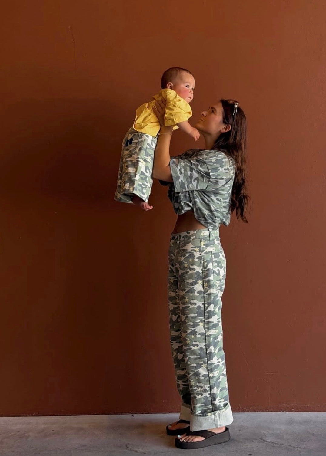 A woman and a baby wearing matching camouflage print clothing, standing against a solid brown wall. The woman is holding the baby up, and both are wearing a matching camouflage long-sleeve crop top and wide-leg pants, with the baby also wearing a matching yellow top.