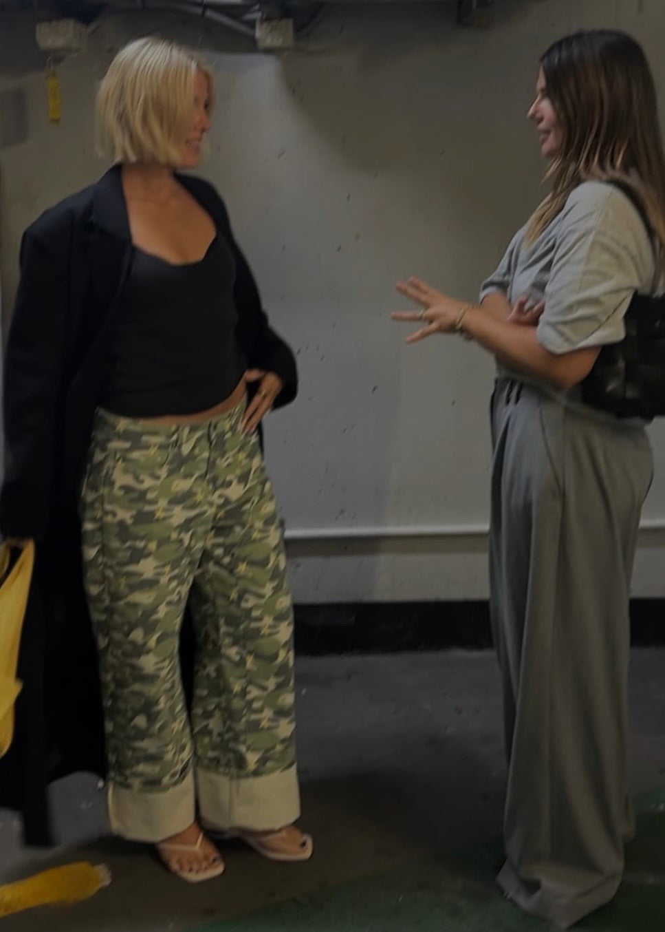 Two women standing and talking in a dimly lit indoor space. The woman on the left wears a black blazer over a black top and wide-leg camouflage pants with rolled cuffs.