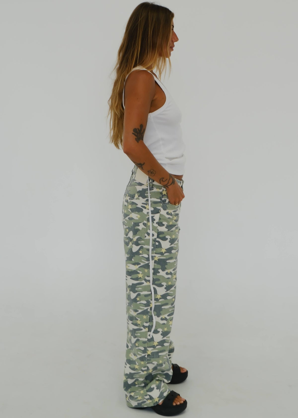 A woman shown in profile wearing camouflage wide-leg sweatpants, a white tank top, and black slide sandals against a plain light gray studio background.