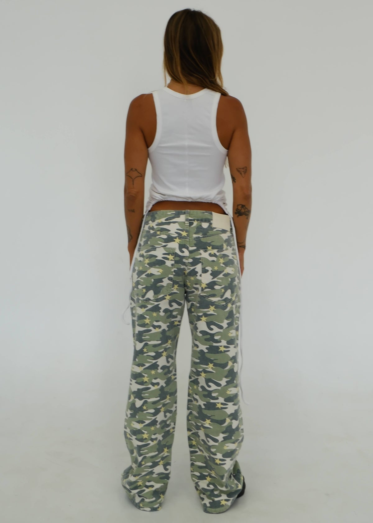 A woman viewed from behind wearing camouflage wide-leg sweatpants, a white ribbed tank top, and black sneakers against a plain white studio background.