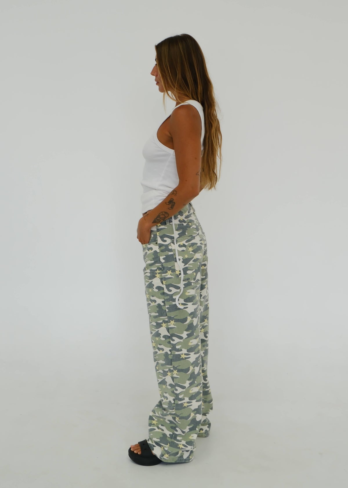 A woman stands in profile wearing camouflage sweatpants, a white tank top, and black slide sandals against a plain white studio background.