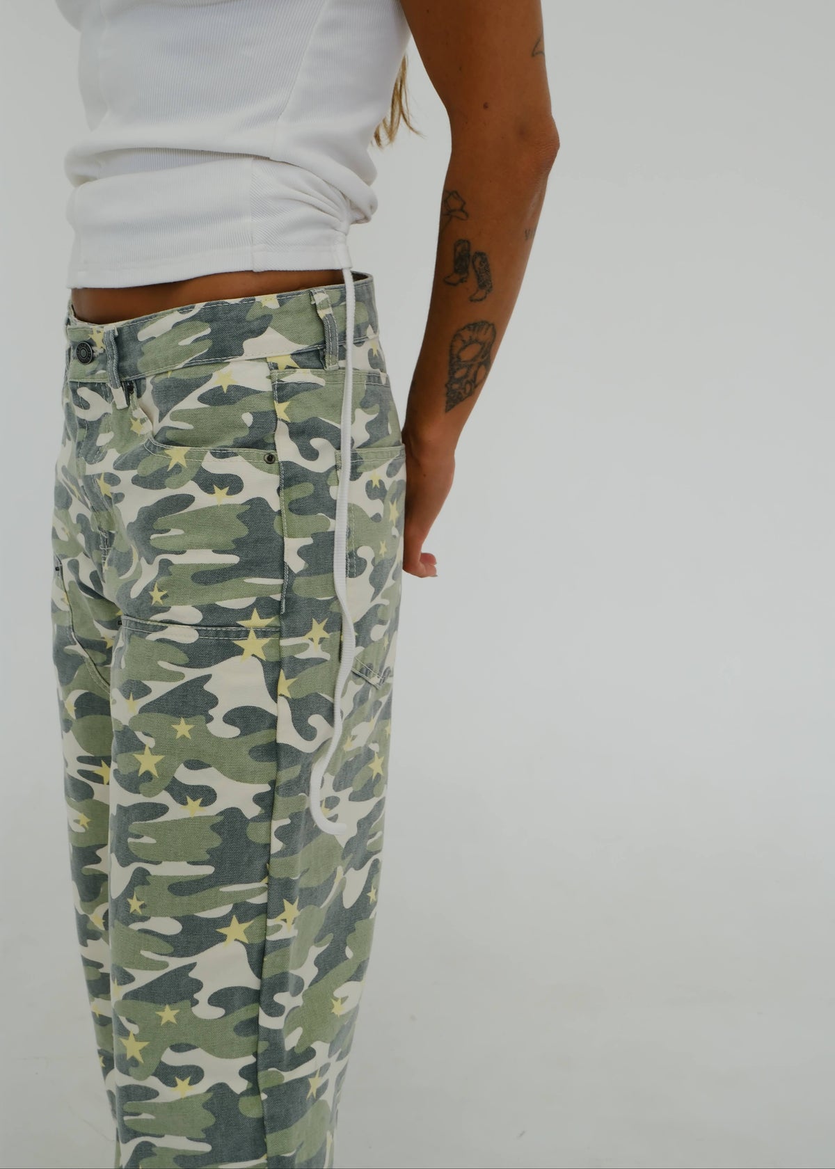 A person wearing camouflage pants with a white side stripe and white drawstrings, paired with a white sleeveless top, shown from the waist down against a plain light gray background.