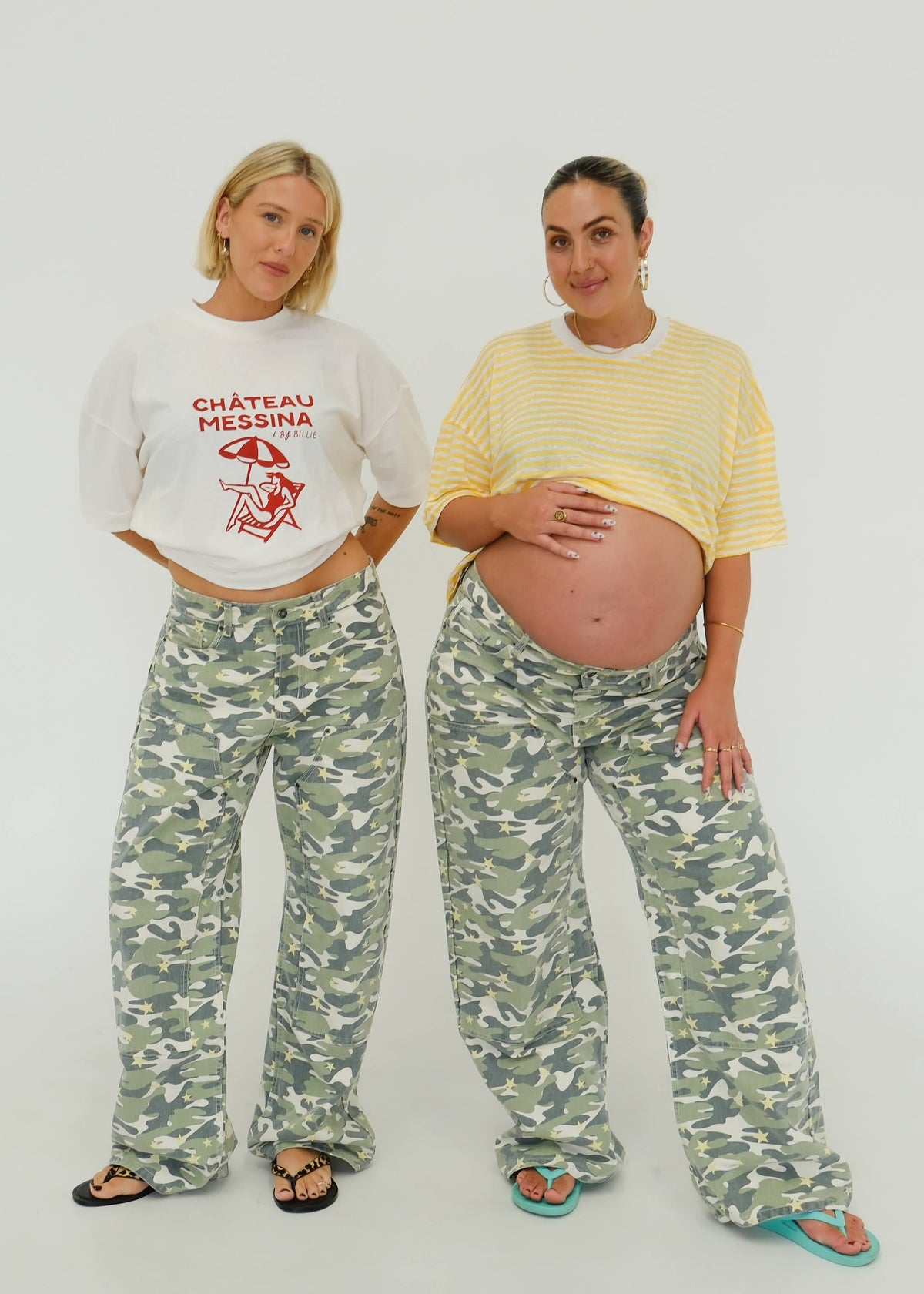 Two women standing against a plain white studio background, each wearing matching camouflage pants and different top styles: one in a white cropped T-shirt with red text and a star graphic, and one in a yellow striped cropped T-shirt with a raised belly.