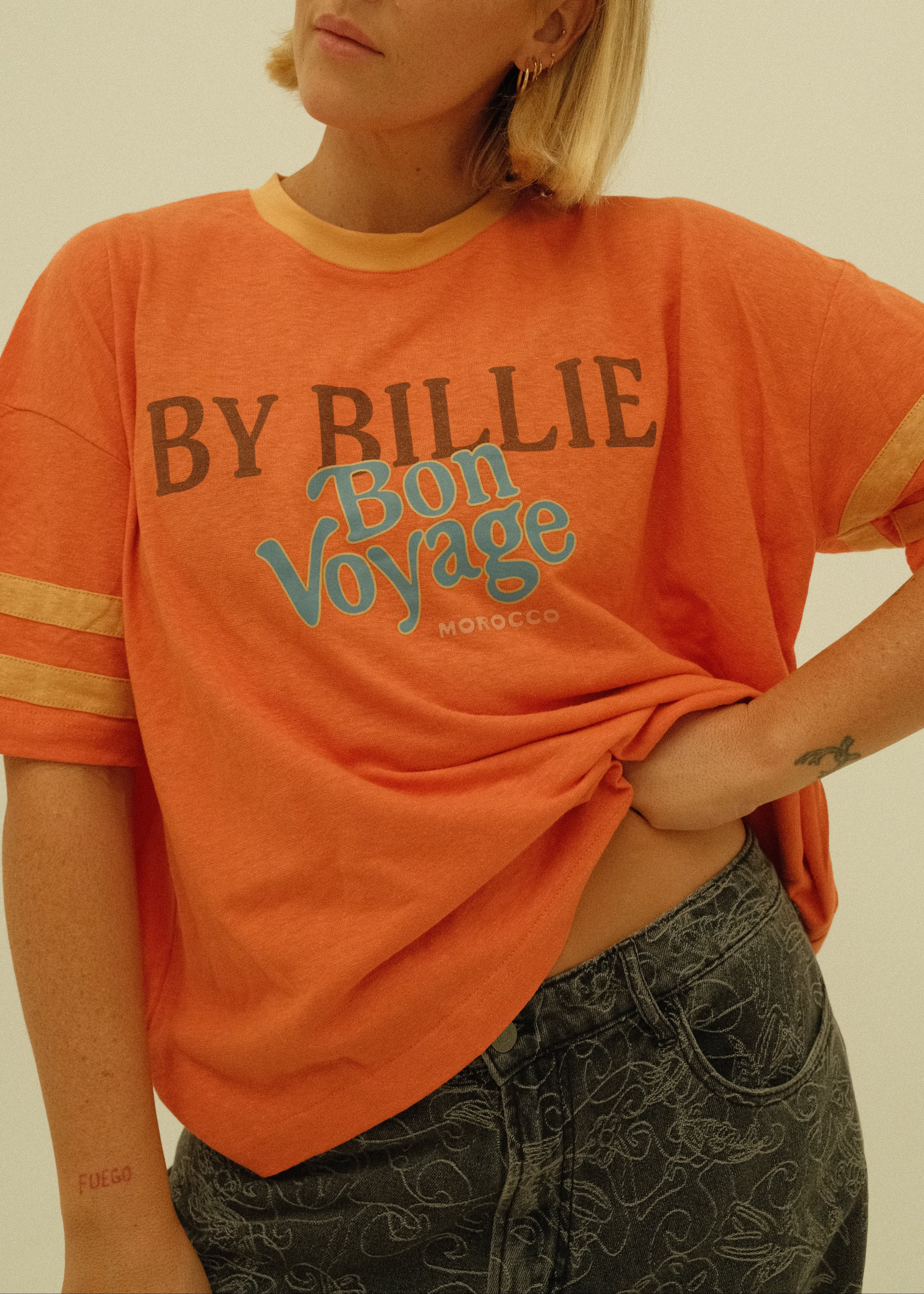 A woman wearing a bright orange graphic t-shirt with 'BY BILLIE Bon Voyage' printed on the front, paired with dark patterned jeans, photographed against a plain off-white background.
