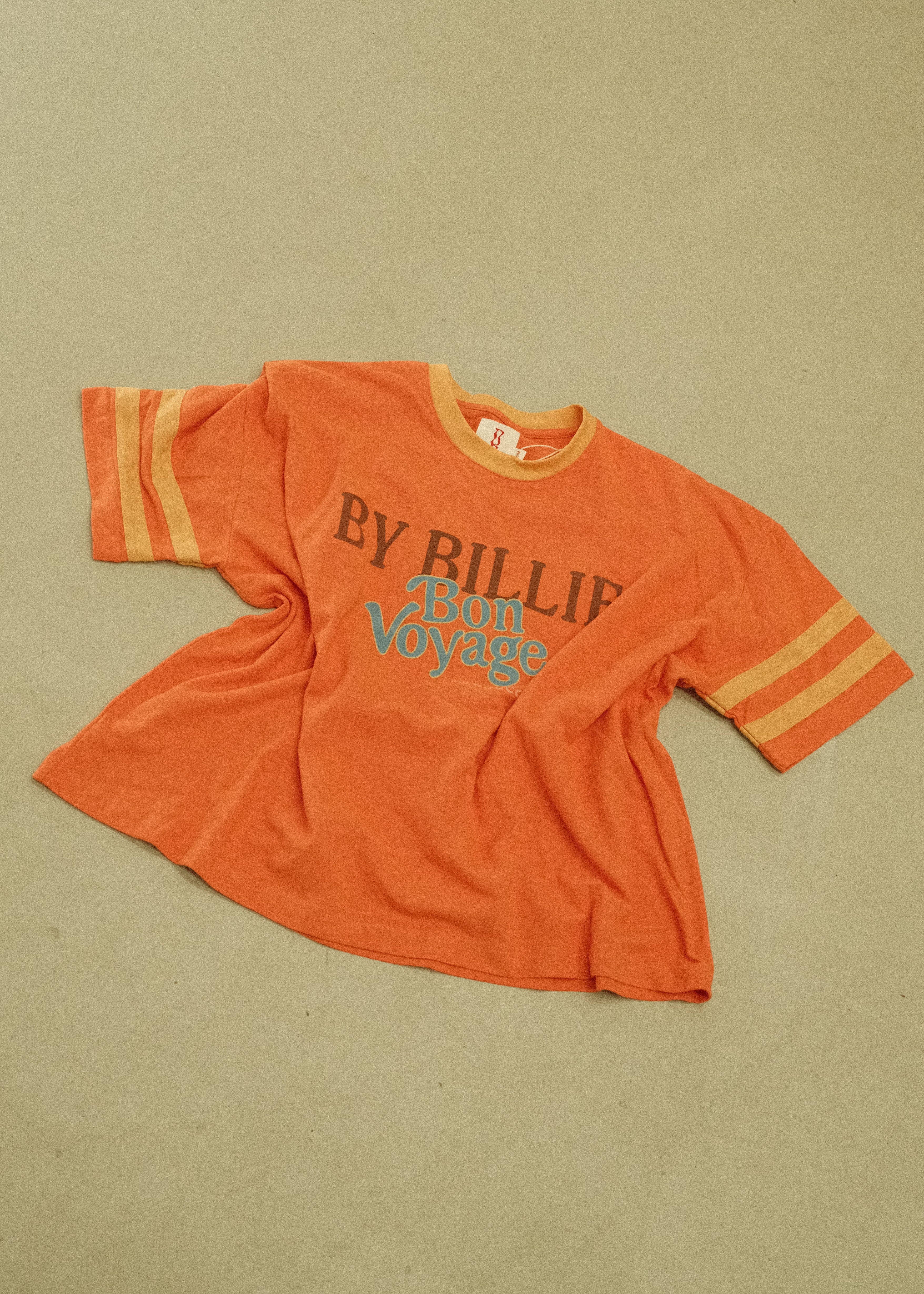 An orange vintage short-sleeve t-shirt with yellow stripes on the sleeves and a blue graphic print reading 'BY RILLIE' on the front, laid flat on a beige background.