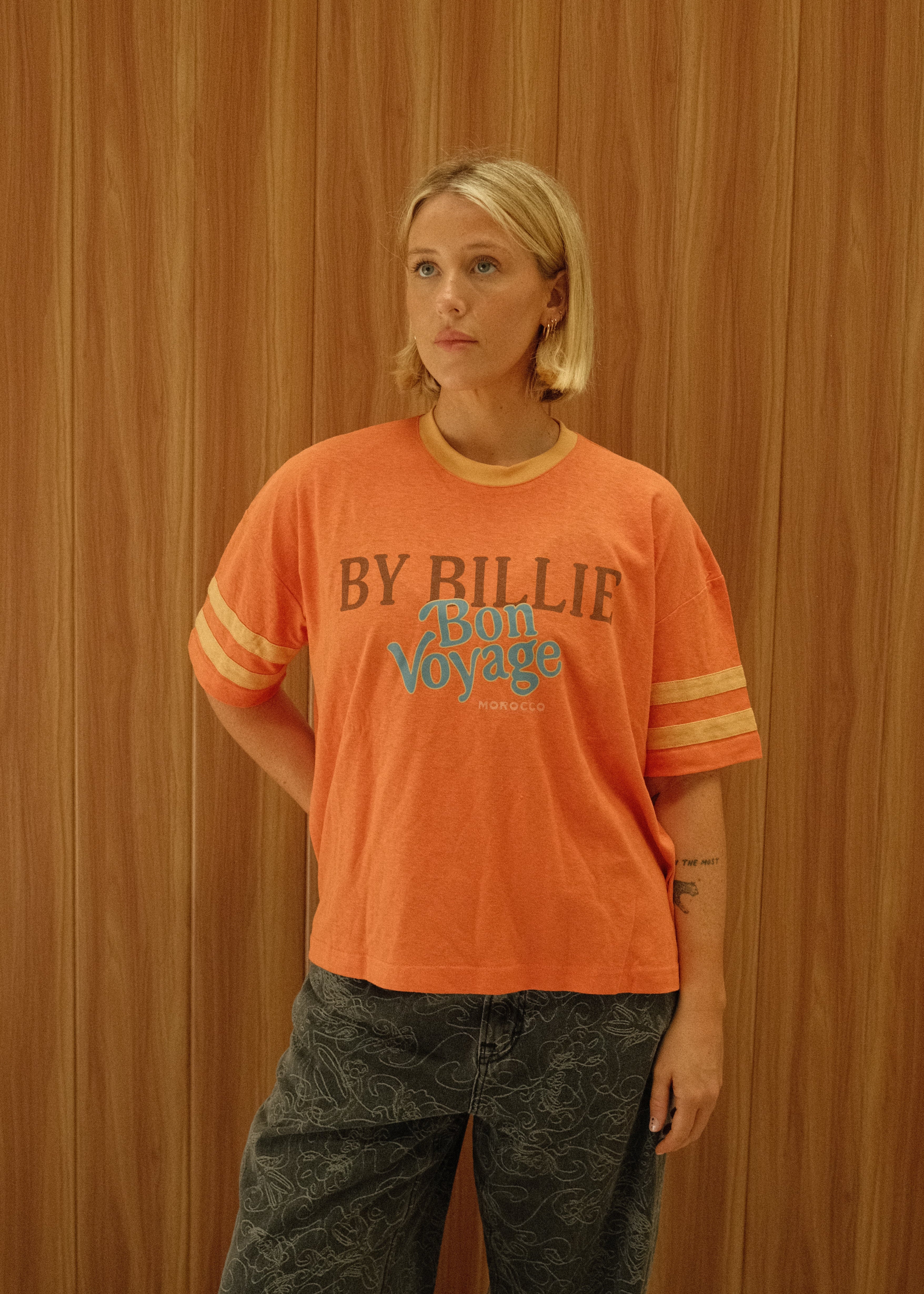 A person lying on a wooden floor wearing an orange short-sleeve graphic T-shirt with yellow stripes on the sleeves and dark gray patterned pants. The shirt has blue and black text reading 'BY BILLIE EASON'.