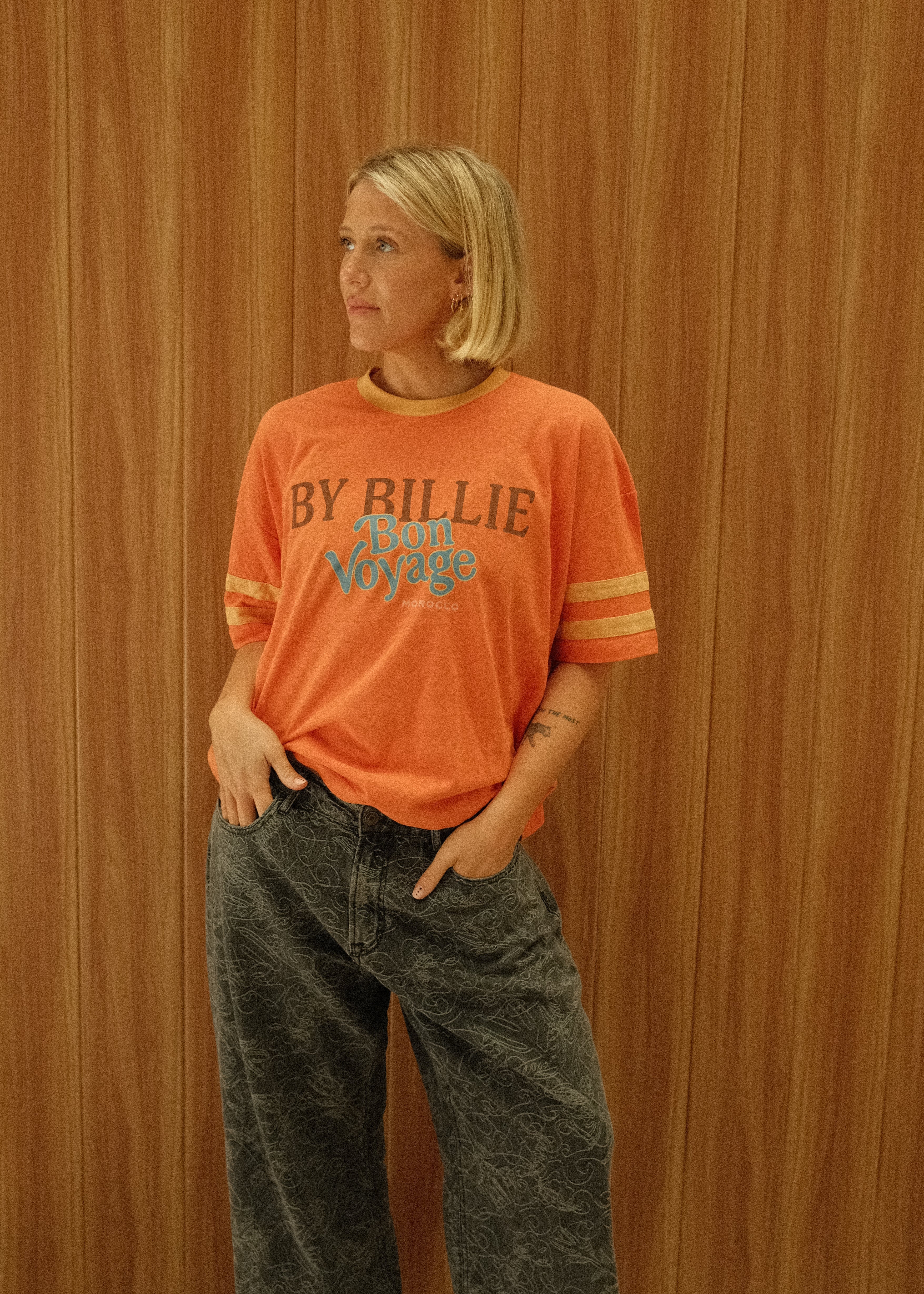 A woman modeling an orange short-sleeve t-shirt with 'BY RILLIE' printed on the front, paired with dark gray jeans and black sandals, standing against a wood-paneled wall.