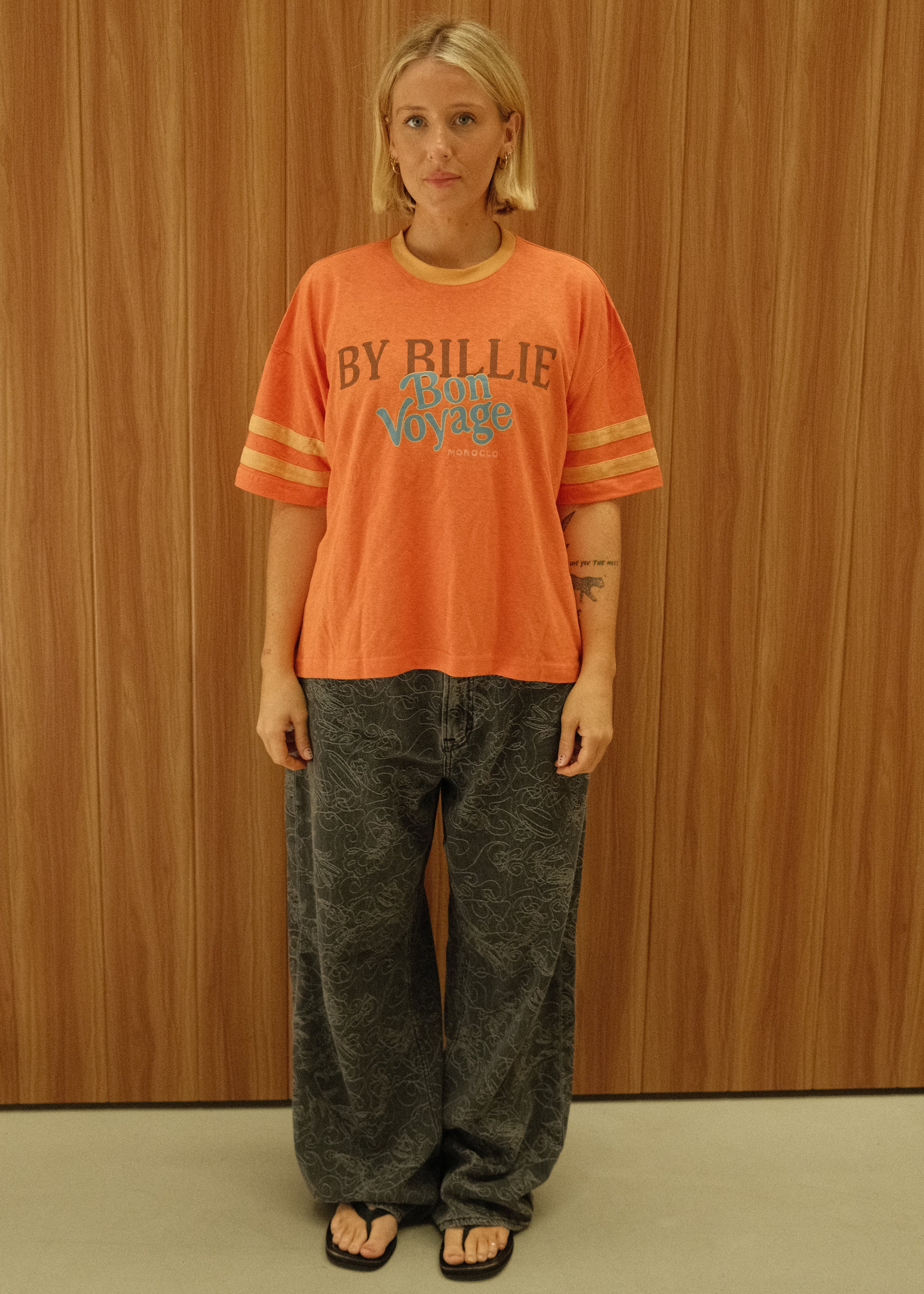 A person standing against a wood-paneled wall wearing a bright orange short-sleeve t-shirt with 'BY BILLIE ELLIS' printed on the front, paired with dark gray jeans and black sandals.