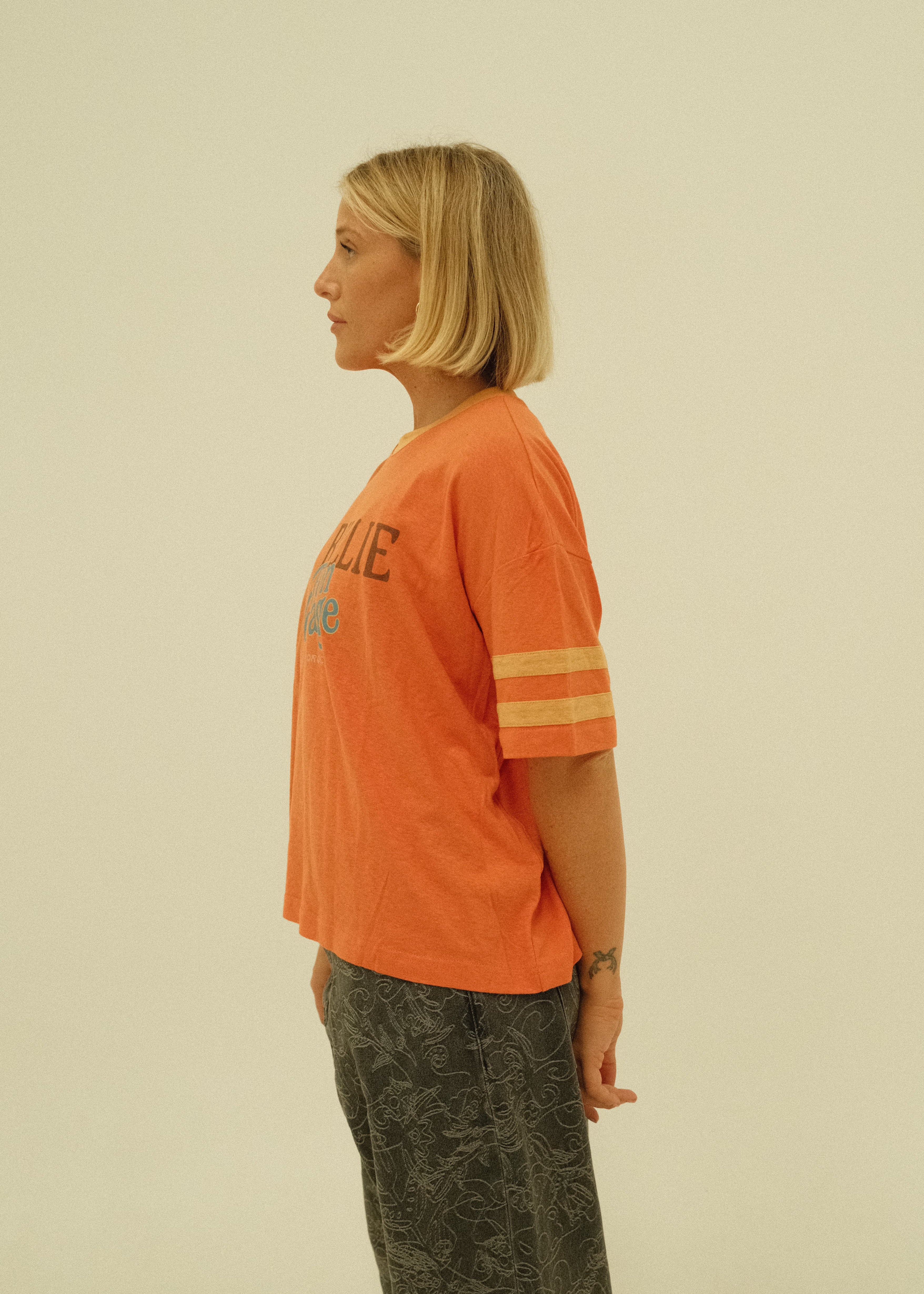 A woman with blonde hair is shown in profile wearing an orange t-shirt with yellow stripes on the sleeve and dark gray pants, set against a solid light beige background.