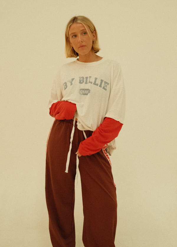 A woman wearing a white graphic t-shirt with red sleeves, paired with brown sweatpants, photographed against a plain off-white background.