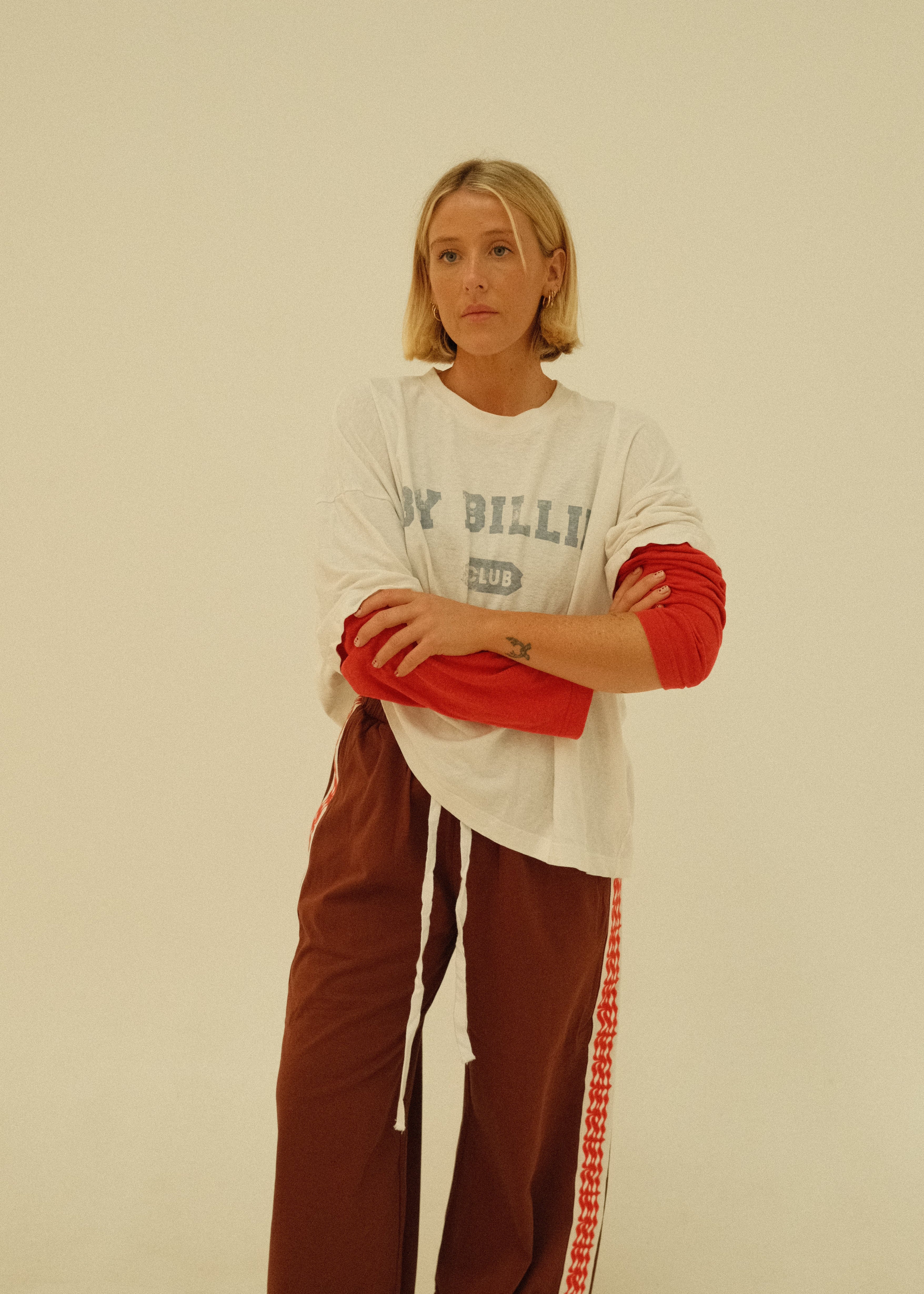 A woman modeling a white t-shirt with red 3/4 sleeves and a gray graphic print, paired with brown sweatpants featuring a red and white stripe down the side, against a plain off-white background.