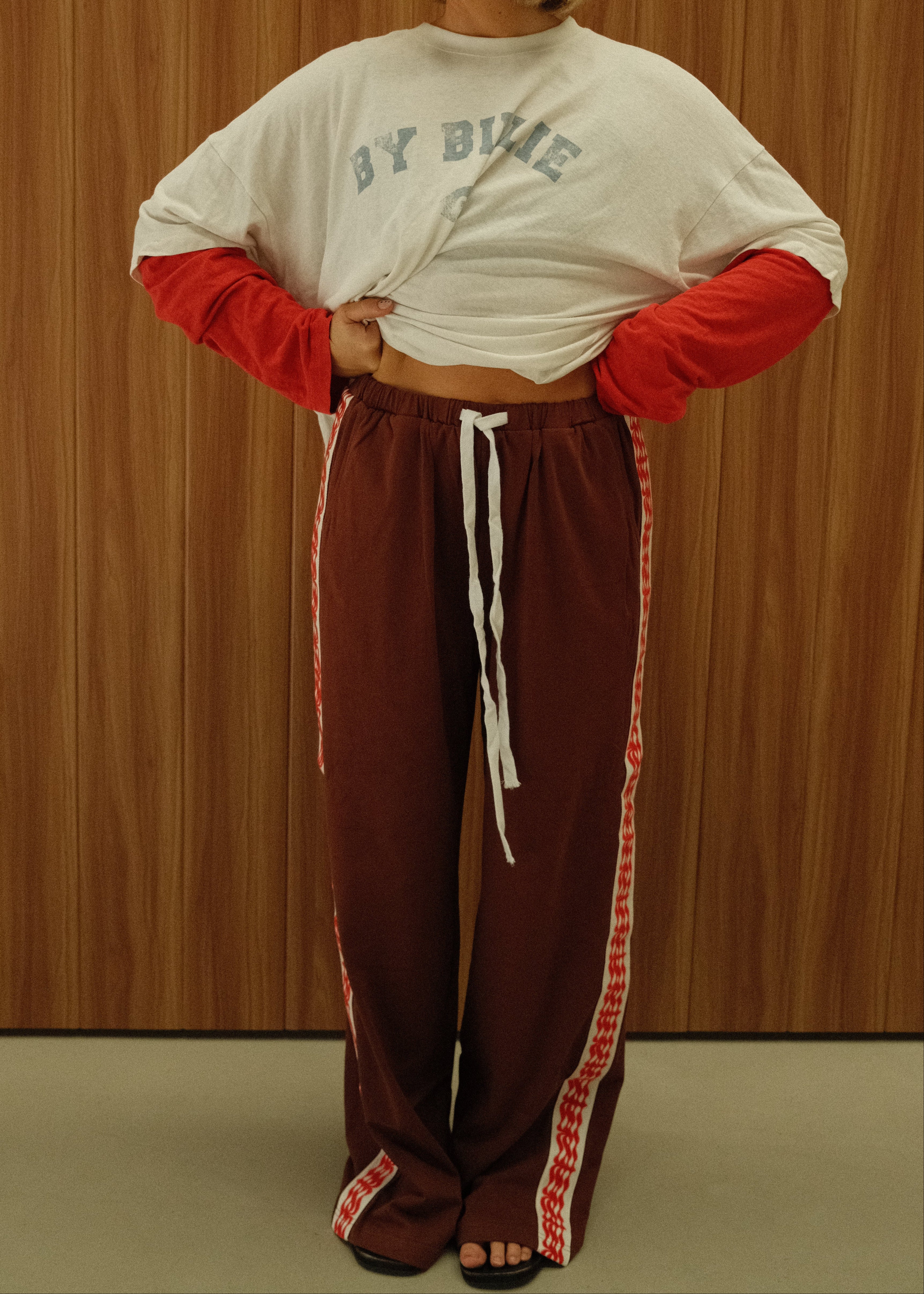 A woman wearing a white t-shirt with red sleeves and matching red-and-white striped athletic pants, standing against a wood-paneled wall.