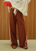 A person wearing a white and red cropped t-shirt with red sleeves, brown wide-leg sweatpants with red and white side stripes, and black slide sandals, standing against a wood-paneled wall.