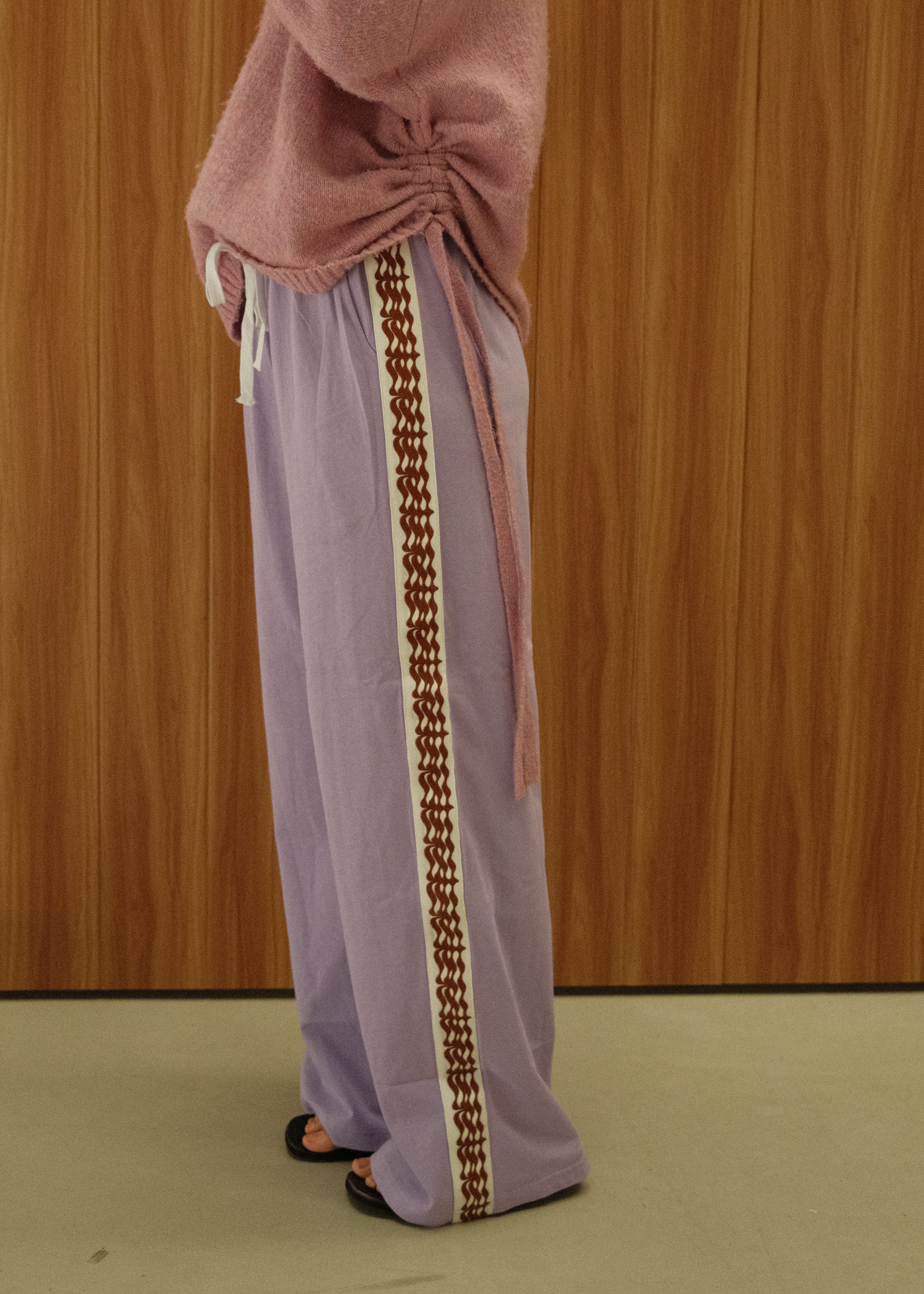 A person wearing a lavender and pink traditional Japanese kimono robe with a brown patterned border, standing against a wooden wall.