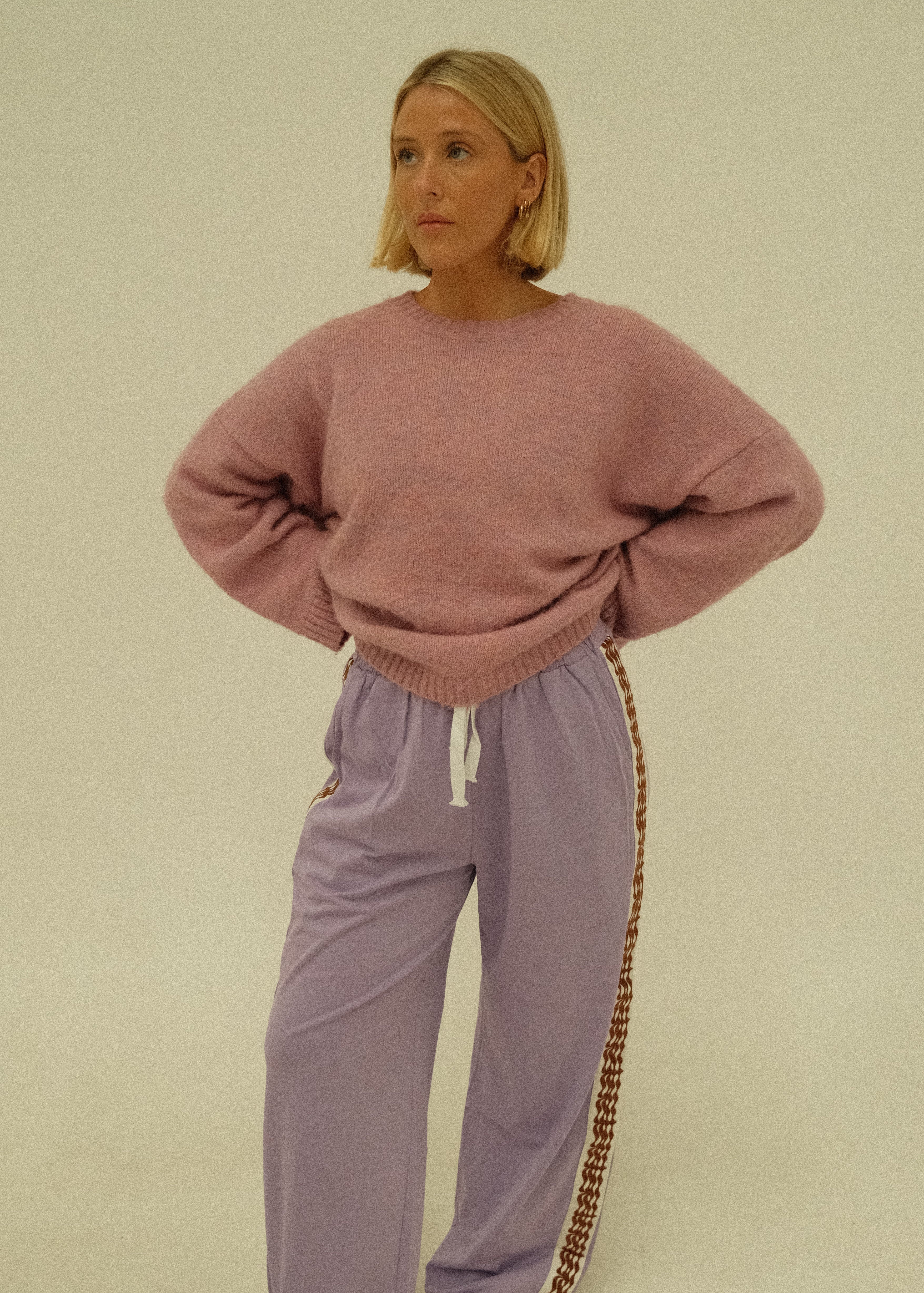 A woman wearing a dusty pink knit sweater and matching lavender wide-leg pants with a brown patterned side stripe, standing against a plain light beige background.