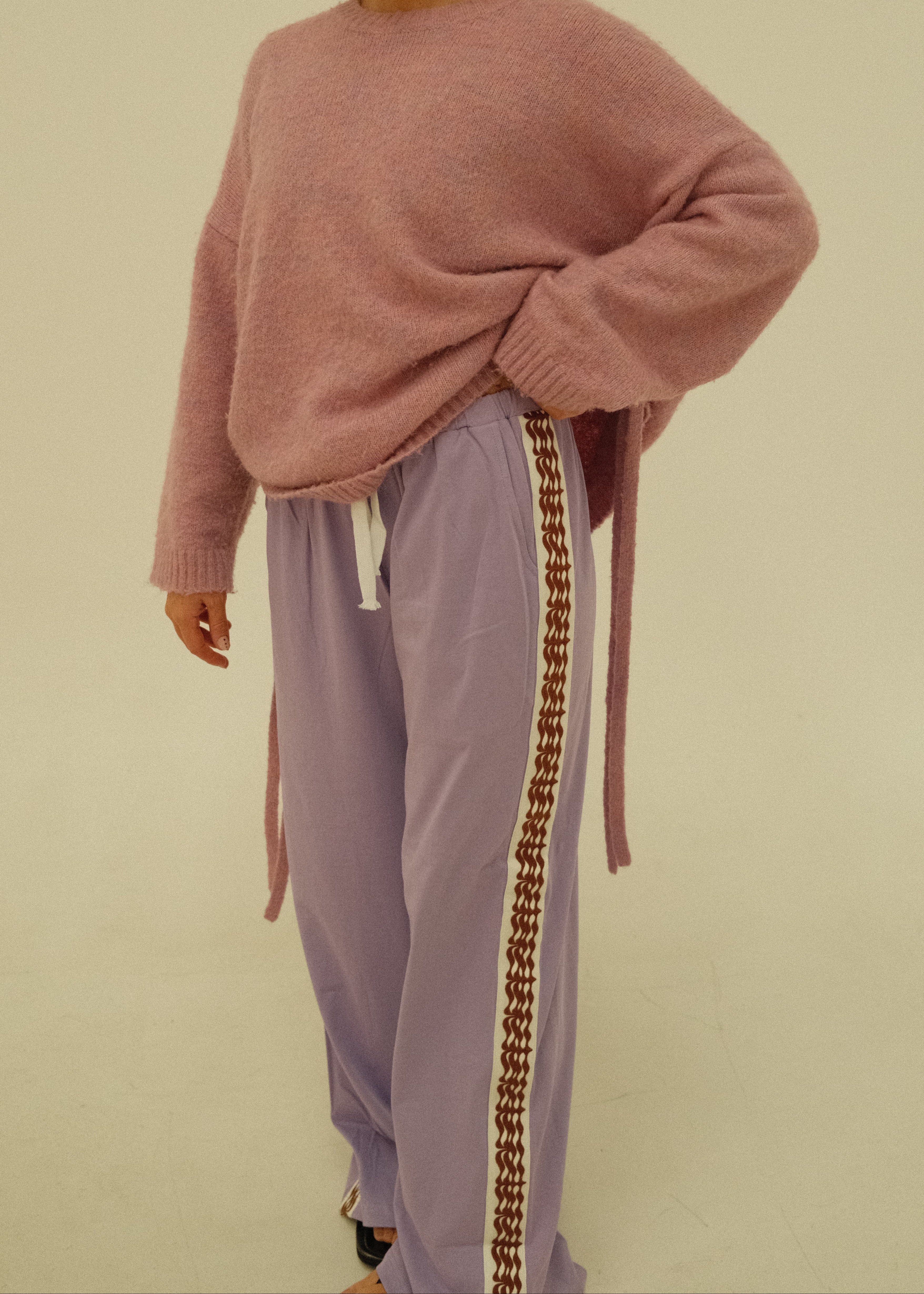 A woman wearing a dusty pink knit sweater and lavender sweatpants with a brown geometric stripe detail, posed against a plain beige background.