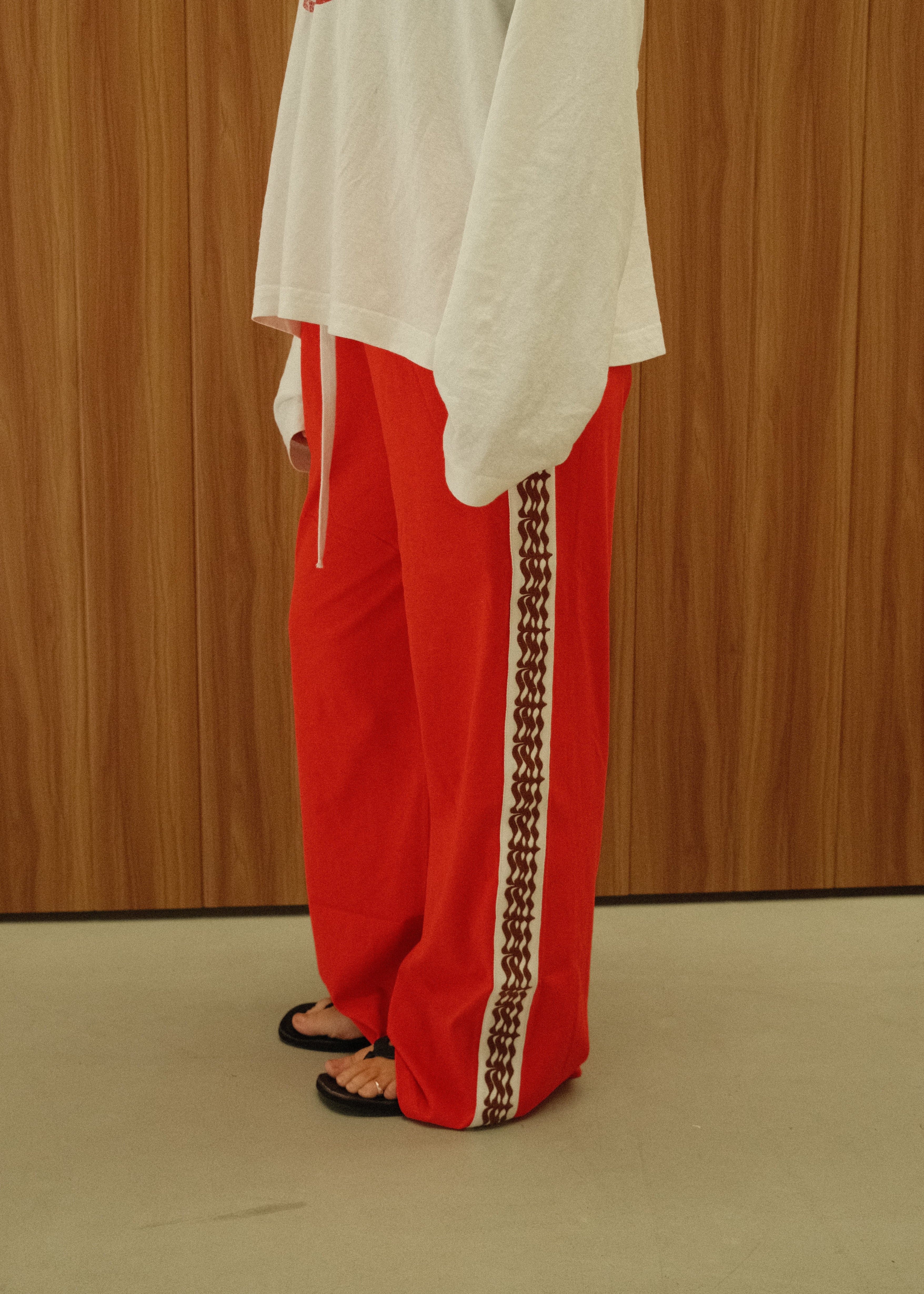 A person wearing a white oversized long-sleeve shirt with red collegiate lettering and bright red drawstring pants with a black-and-white patterned side stripe, standing against a wood-paneled wall.