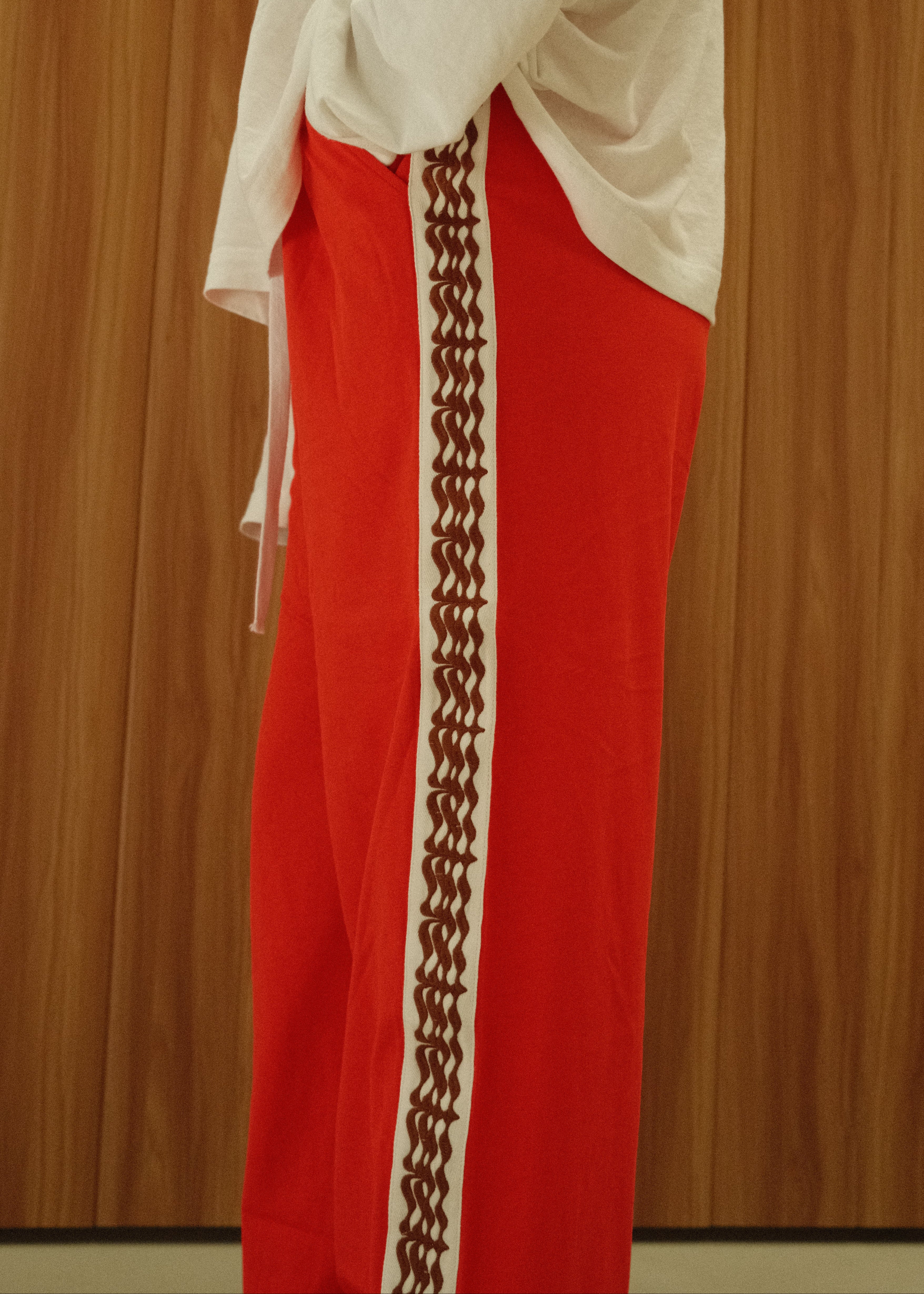 A person wearing bright red track pants with a wide white side stripe featuring a brown geometric pattern, paired with a loose white top, standing against a wood-paneled wall.