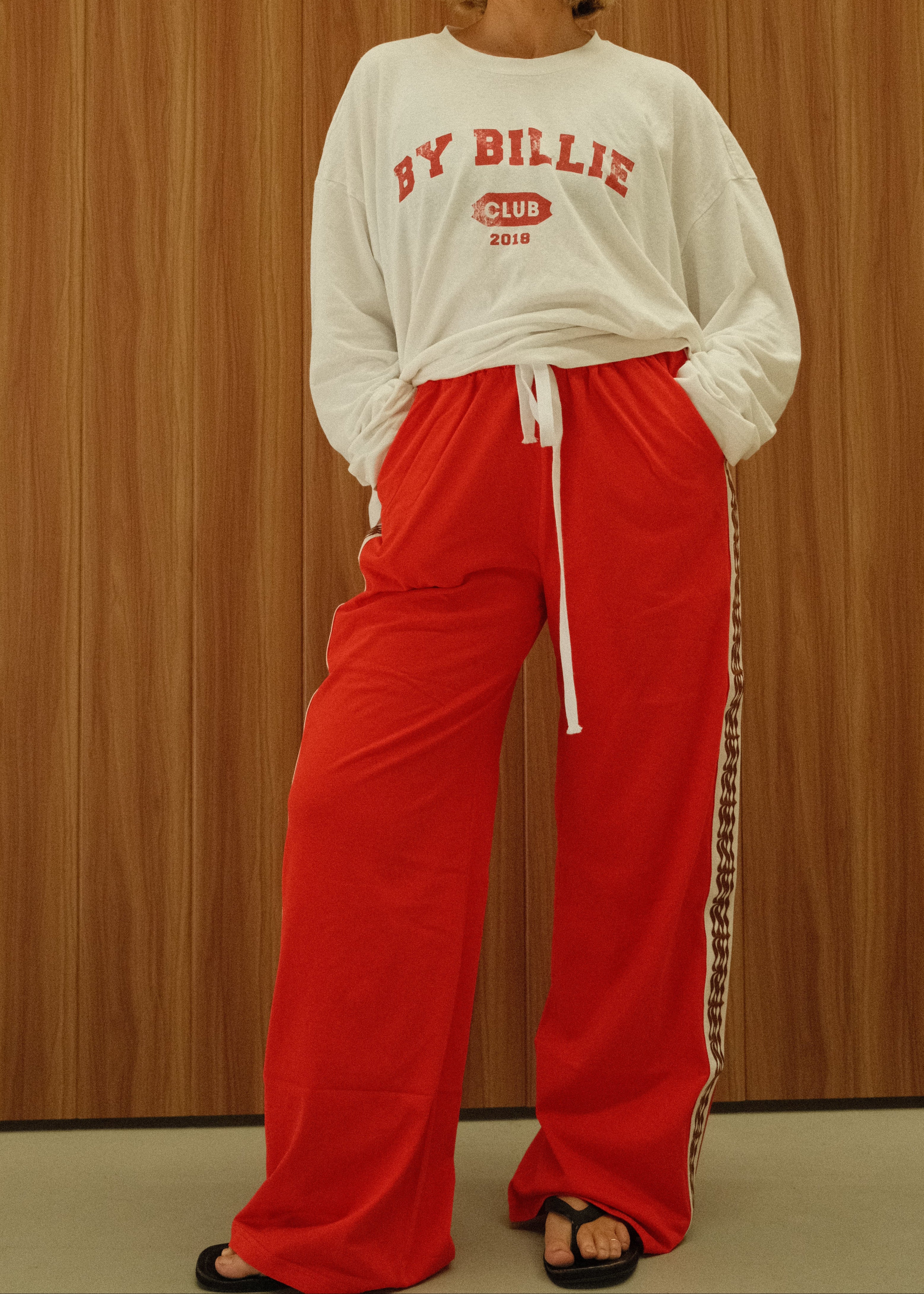 A woman stands against a wood-paneled wall wearing a white sweatshirt with red collegiate-style text reading 'BY BILLIE CLUB EST. 2019' and bright red wide-leg sweatpants with a white drawstring and a black-and-white patterned side stripe.