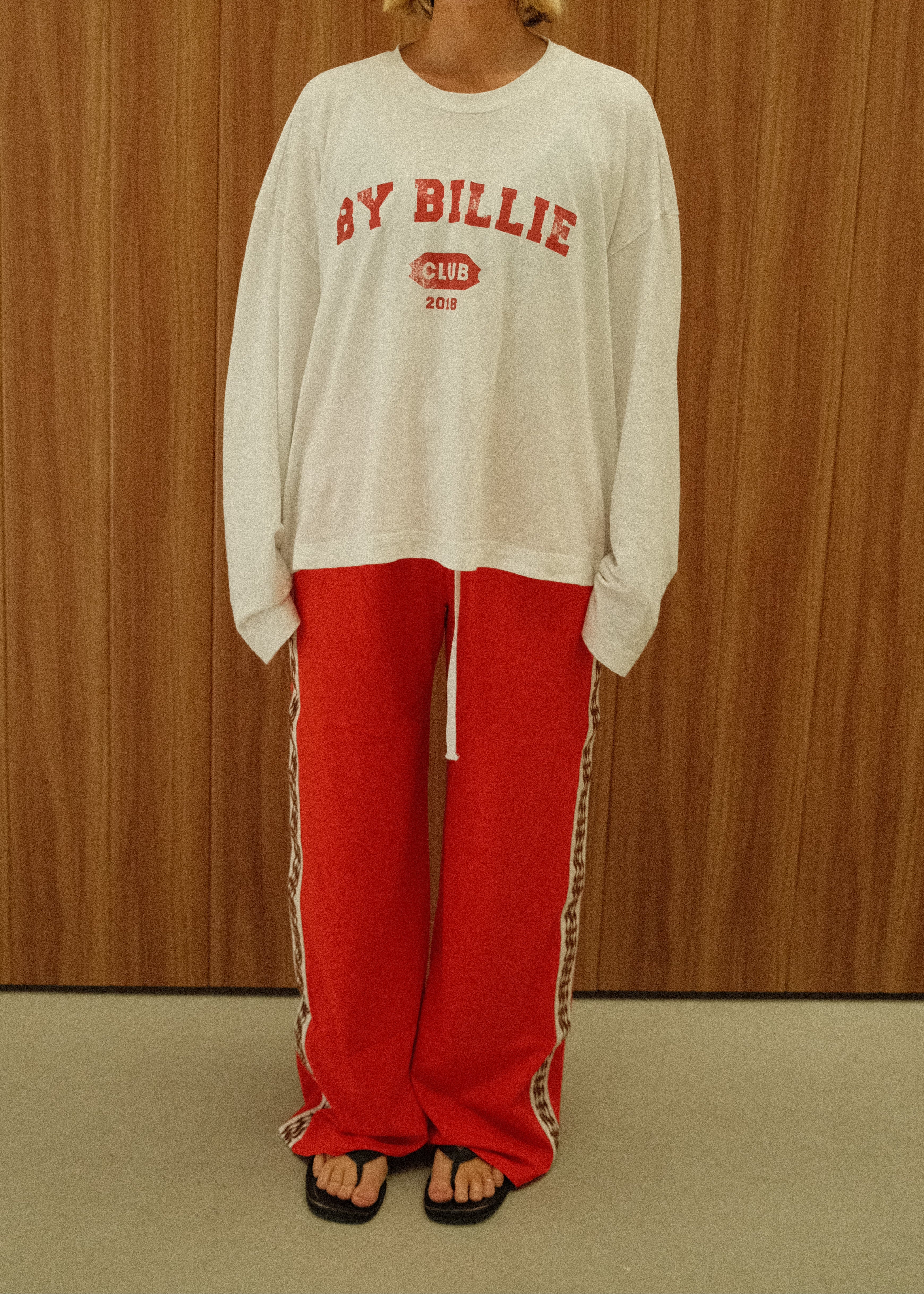 A woman stands against a wood-paneled wall wearing an oversized white long-sleeve shirt with red collegiate-style text reading 'BY BILLIE CLUB' and bright red wide-leg pants with white side stripes.